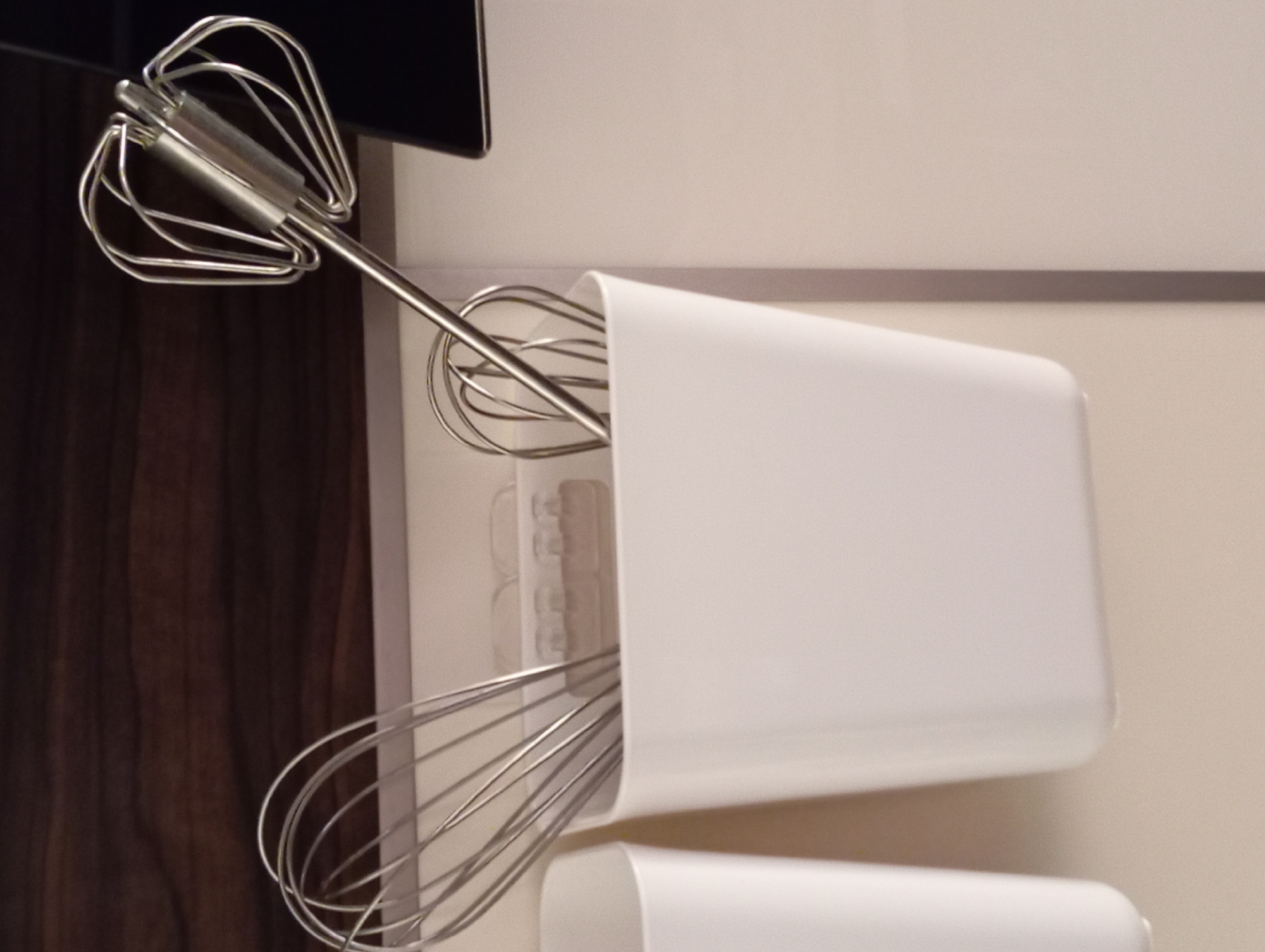 The image shows a close-up of two metal whisks, likely for a hand mixer, stored in a white container. The container is cylindrical and open at the top. One whisk is almost entirely contained within the holder, with only its handle and some of the wire loops visible. The second whisk is positioned above the container, with its wire loops extending outward, showcasing its intricate design. The handle of this upper whisk is metallic and appears to have some cylindrical attachments near its base.
The background of the image is divided into two distinct areas. To the left, a dark, vertically paneled surface suggests wooden cabinetry or a dark wooden door. To the right, a lighter, smooth surface, likely a wall or cabinet door, is visible. A thin horizontal stripe, possibly a metallic trim or a seam, runs across this lighter surface. The lighting appears to be artificial and even, with no strong shadows, suggesting an indoor setting.
No people are visible in the image, and the activity is implied to be storage of kitchen utensils. The setting appears to be a kitchen or a utility area. There is no visible text in the image. The overall impression is one of neat organization of kitchen tools. The location context of Baden, Austria, does not provide specific visual cues within the image itself.