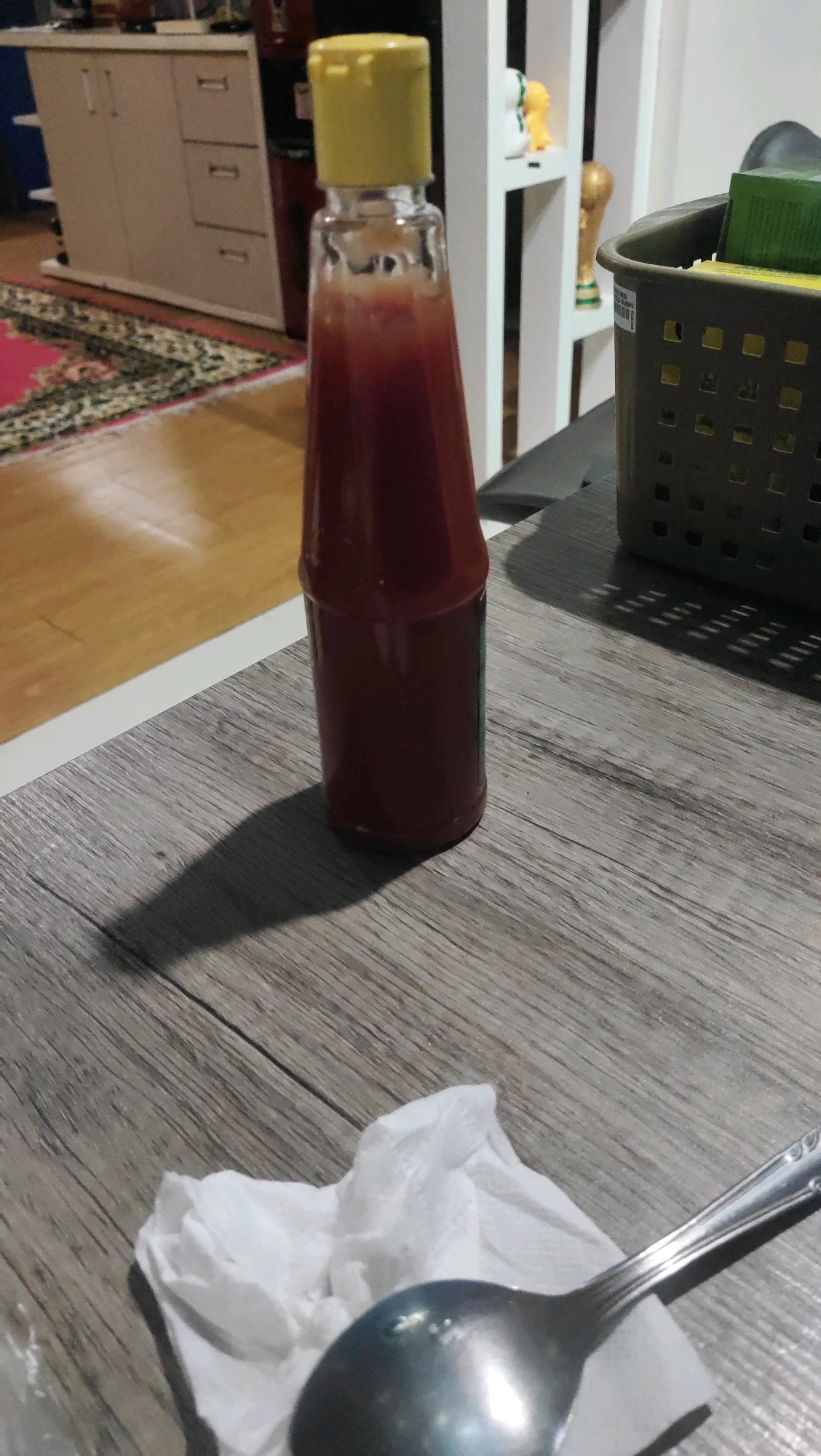 A bottle of red sauce with a yellow cap stands on a wooden table, casting a shadow. To the right, a dark basket holds some items, one of which is green. In the foreground, a spoon rests on a crumpled white napkin. In the background, a cabinet with drawers and a colorful rug are visible. The environment appears to be indoors, possibly a dining area.