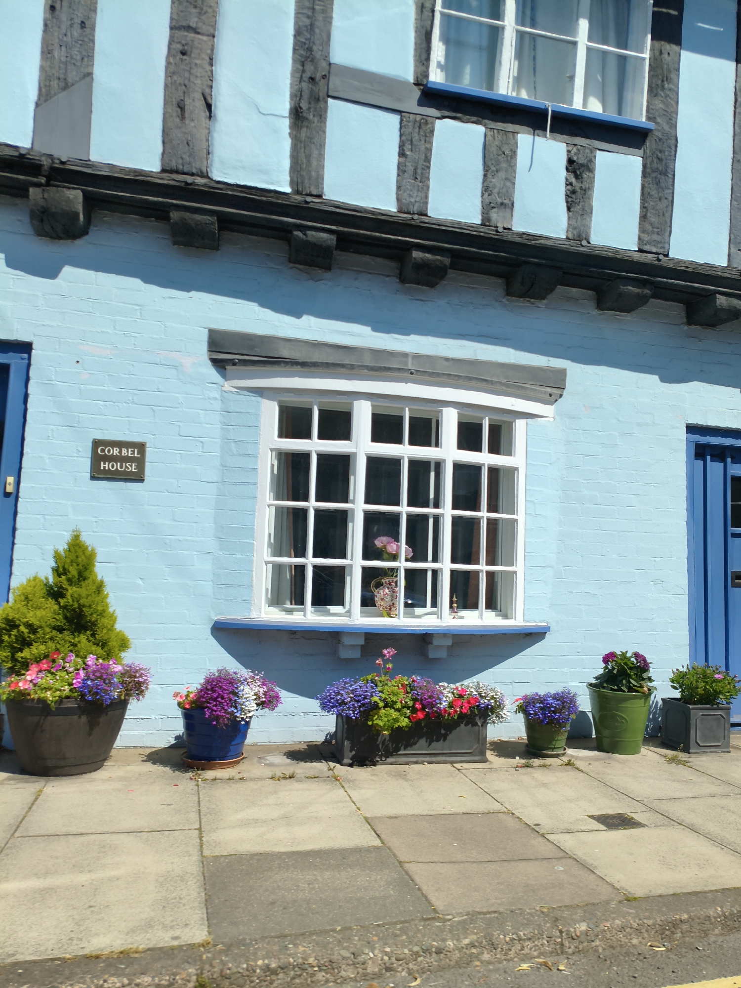 The image shows the exterior of Corbel House, a building located in Ludlow, United Kingdom. The house is painted a light blue and features traditional timber framing with dark wooden beams. A large white-framed window with multiple panes is visible in the center of the facade. A small rectangular sign with "CORBEL HOUSE" written on it is mounted on the wall to the left of the window. Several potted plants with colorful flowers are arranged on the paved sidewalk in front of the house. The sky is clear and the scene appears to be illuminated by sunlight.