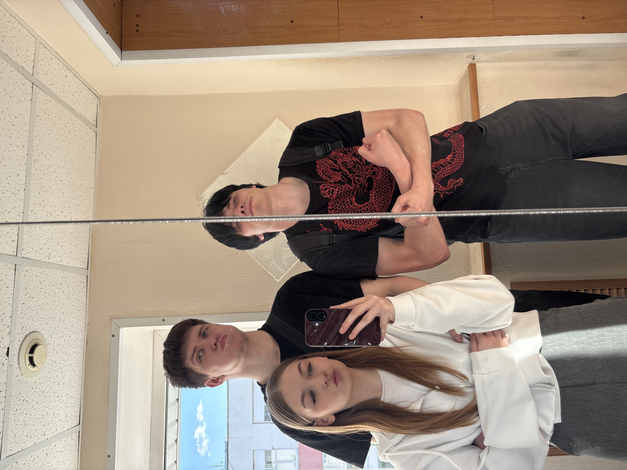 A selfie of three people is captured in a mirror indoors. The reflection shows a woman and two men. The woman is holding the phone and wearing a white hoodie, the man next to her is wearing a black shirt. In the reflection, the third man is seen wearing a black shirt with a red dragon design. The location appears to be inside a building, indicated by the ceiling tiles and the window in the background. A glimpse of a blue sky and building visible through the window suggests the setting may be the city of Chaikovsky, Russia.