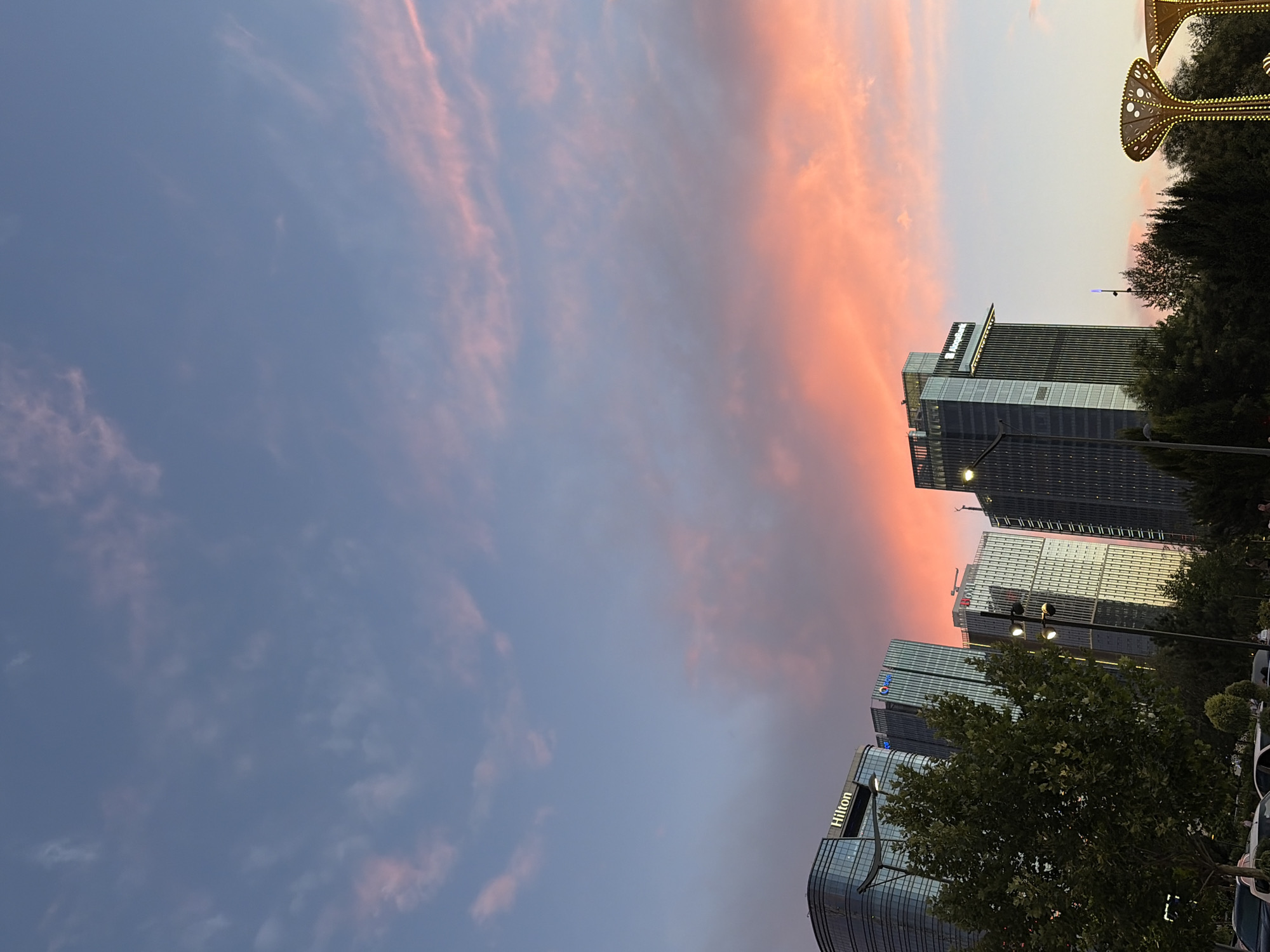 The image depicts a cityscape with towering modern buildings against a dramatic sky. The sky is a gradient of colors, transitioning from a deep blue on the left to vibrant hues of pink and orange on the right, suggesting a sunset or sunrise. Wispy clouds are scattered across the sky, catching the warm light.
Several contemporary buildings are visible, characterized by their glass facades and sleek designs. One prominent building displays the "Hilton" logo in illuminated letters. The architecture suggests an urban, possibly business district setting. Lush green trees are interspersed throughout the scene, adding a natural element to the man-made structures. In the upper right corner, a decorative structure with a golden, illuminated design is partially visible, hinting at a festive or entertainment area.
The scene appears tranquil and visually striking due to the contrast between the natural sky phenomena and the urban environment. The lighting suggests it is either early morning or late evening. No people are clearly identifiable in the image, but the presence of cars on the right side indicates a public area or street. The overall impression is one of a serene yet dynamic urban landscape bathed in the warm glow of twilight.