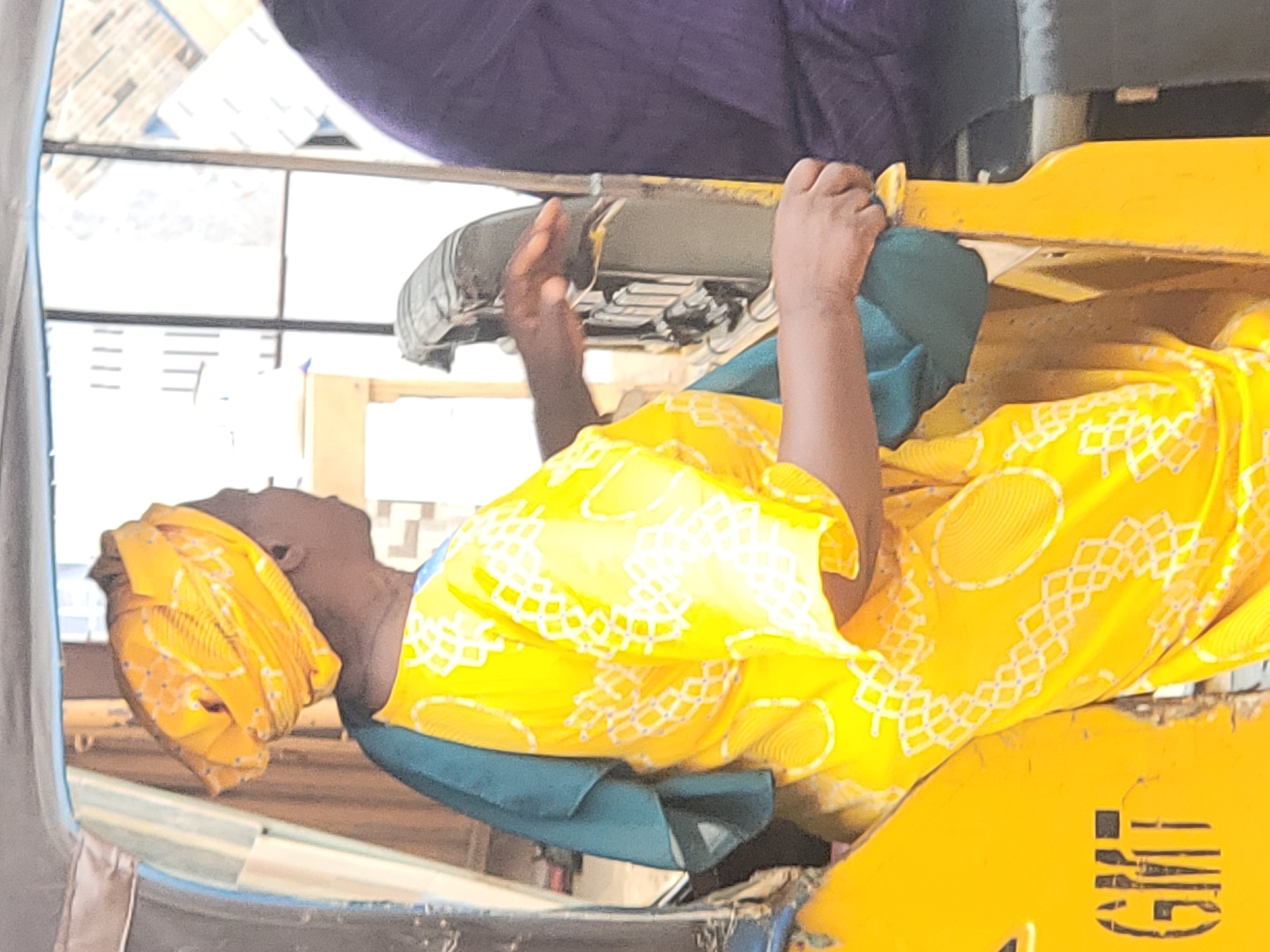 This image, captured in Maiburiji, Nigeria, depicts a person seated or positioned within an open-sided vehicle, likely a tricycle or similar local transport. The perspective is tilted, showing the person horizontally across the frame.
The subject has dark skin and is wearing a striking yellow garment with a white and light blue geometric pattern, possibly a traditional dress or top. A green fabric is visible underneath this main garment. A matching yellow headwrap covers their head. Their hands are raised and grasping a metal frame, which appears to be the backrest of a seat or a structural bar of the vehicle, suggesting they are holding on for stability. The person's face is visible, looking upwards and slightly to the left, with a neutral and attentive expression.
The lighting is bright, indicating it is daytime. In the lower right corner, a yellow metal panel of the vehicle is visible, bearing partially obscured black stenciled text that appears to read "G.M.E." or similar letters. A dark purple fabric or cushion is visible in the upper background. The background beyond the vehicle shows blurred light-colored structures, possibly buildings or interior elements, with horizontal lines. The scene suggests a moment during travel or commute within the local setting.
