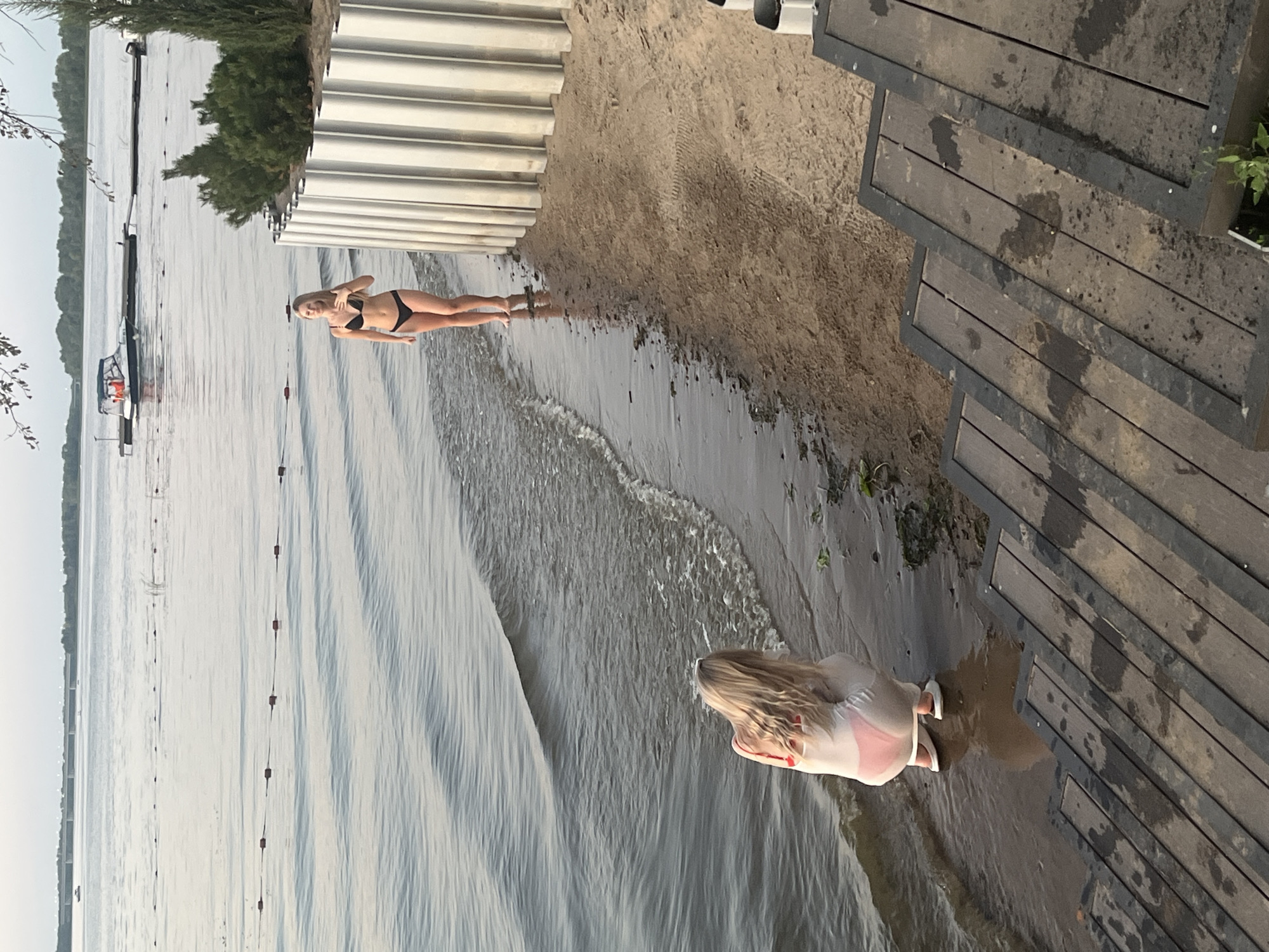 The image captures a serene scene at the Shumeika River embankment in Shumeika, Russia. Two women are present on a sandy beach adjacent to the river. In the foreground, one woman is crouched near the water's edge, wearing a white cover-up over pink bikini bottoms and white slip-on shoes. She appears to be examining something in the shallow water. In the mid-ground, a second woman stands on the beach, wearing a black bikini and facing the river. She has her hand near her chest. The river stretches into the background, with a boat dock and a small boat visible on the water. The beach is demarcated by a metal fence-like structure and wooden stairs lead down to the beach from the right side of the frame. The overall ambiance is one of a leisurely day by the water.