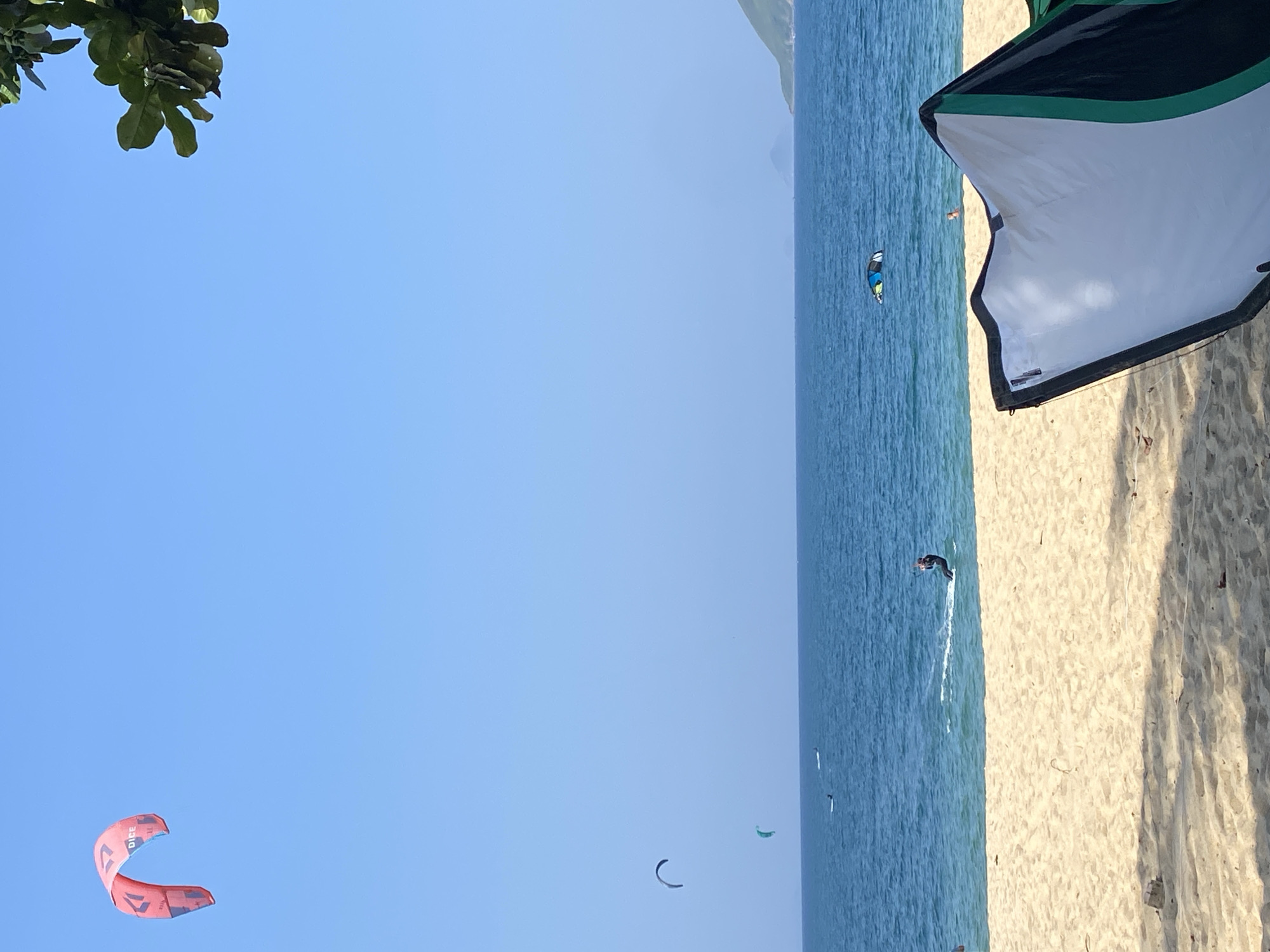 The image depicts a sunny day at a beach in Nha Trang, Vietnam. In the foreground, the sandy beach is partially covered by the shadow of a large, colorful kite, which is predominantly white with black and green accents. Further out, the turquoise ocean stretches towards the horizon, where a hazy coastline is visible.
Several individuals are engaged in water sports. A prominent red and blue kite, labeled "DICE," is high in the clear blue sky. In the water, multiple kitesurfers are active. One kitesurfer, wearing dark attire, is actively riding a board and creating a spray of water. Another two kites, one dark grey and one green, are visible further out at sea, suggesting other kitesurfers are present. A person in the water near the shore appears to be swimming or standing. The overall scene conveys a recreational beach environment with active water sports.