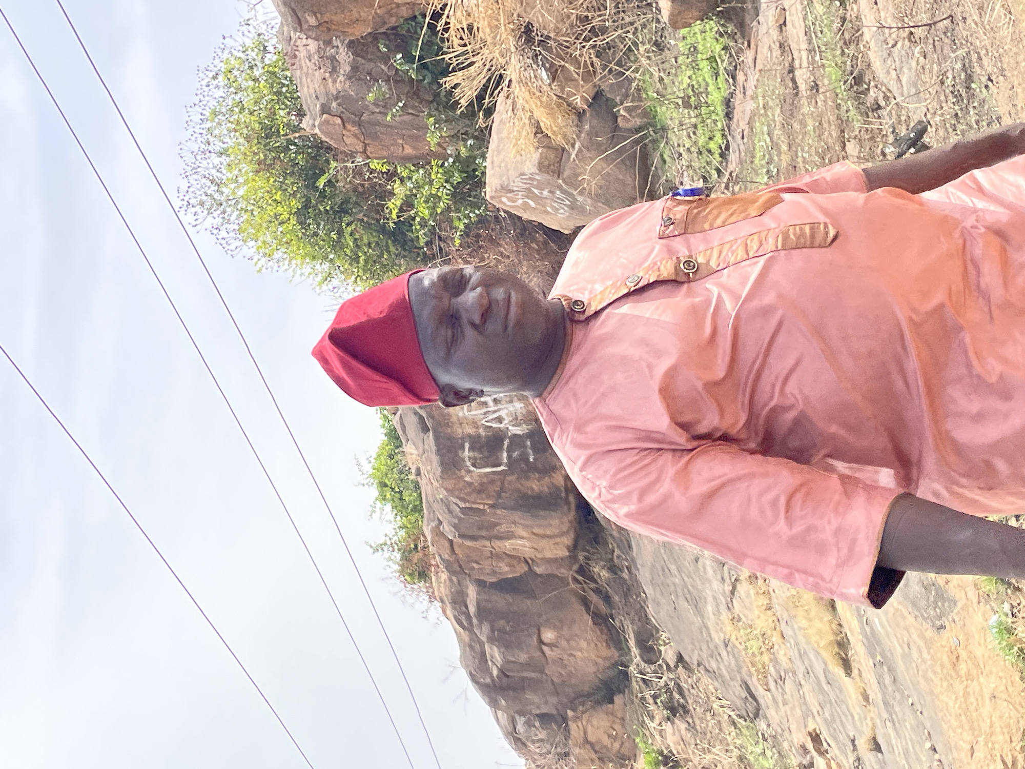 The image features a man wearing a peach-colored garment and a red cap, positioned against a rocky, natural landscape. The background includes vegetation and rock formations, and overhead power lines are visible. The man appears to be outdoors, and based on the prompt, the location is likely in the city of Jos, Nigeria.