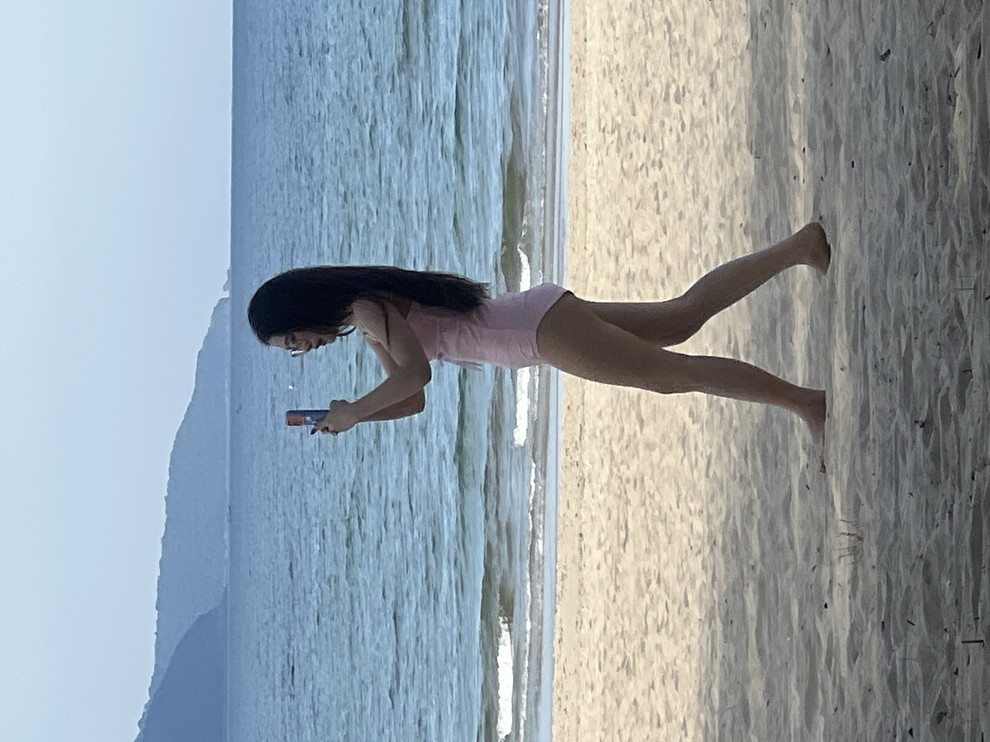 A full-body, side-profile shot captures a young East Asian woman walking on a sandy beach in Đà Nẵng, Vietnam, holding a smartphone in her right hand, seemingly taking a picture. She is wearing a light pink one-piece swimsuit and glasses. Her long, dark hair is loose and falls over her shoulders. The woman is positioned in the mid-ground of the image, with the ocean and a hazy coastline in the background. The sea exhibits a serene blue hue, with gentle waves lapping the shore. The sand is a light beige, with some dried vegetation scattered sparsely. The sky is a pale blue, indicating it could be late afternoon or early morning.