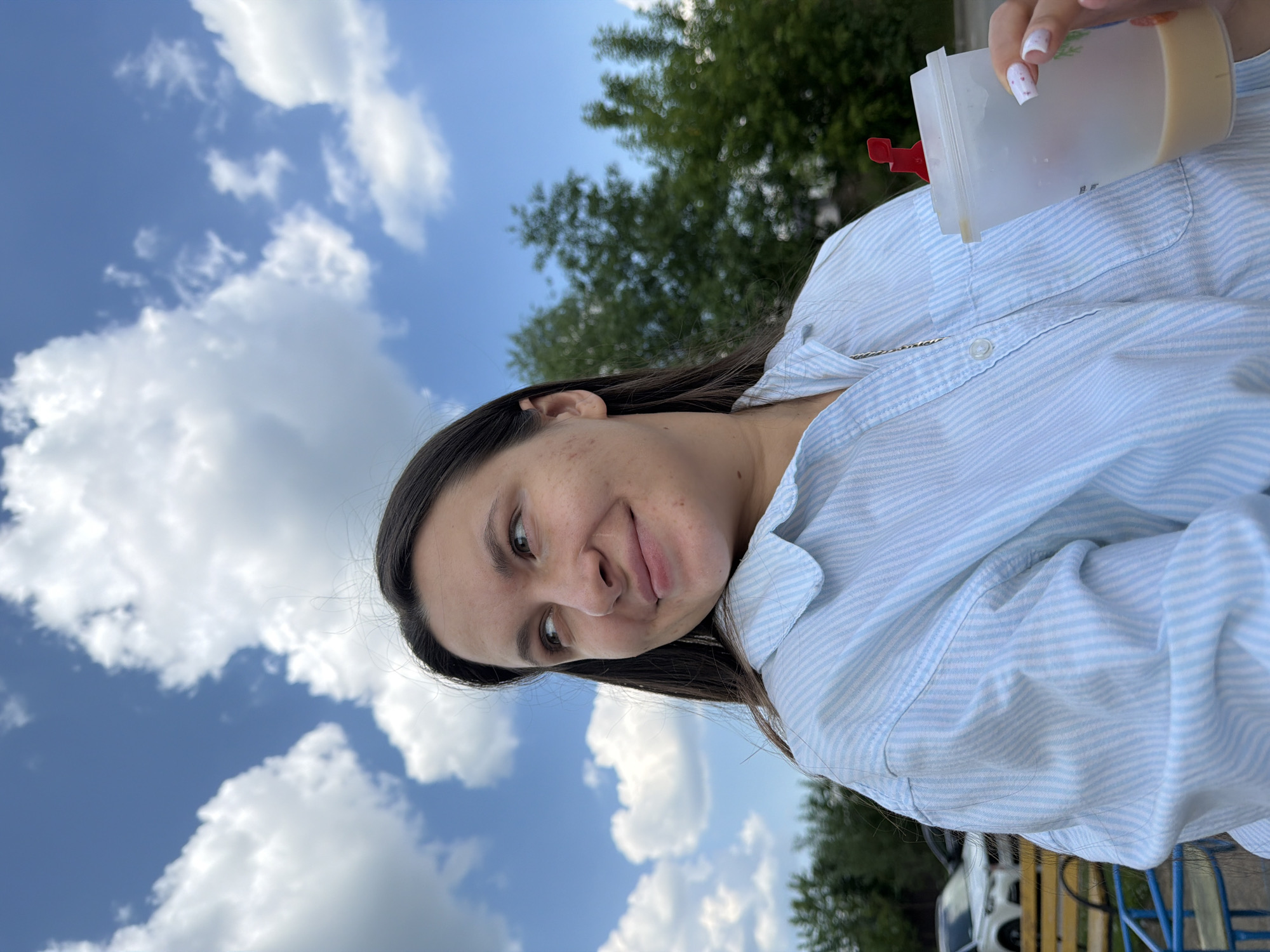 A young woman with long dark hair, wearing a light blue and white striped button-down shirt, looks to her left with a slight smile. She is holding a clear plastic cup with a red lid and straw in her right hand. The background is a blue sky with fluffy white clouds, and some green trees. In the lower left corner, a yellow park bench and blue railing are visible, along with a white car parked in the distance. The image appears to be taken outdoors during the daytime. The location is identified as Akulovo (Gp Kubinka), Russia.