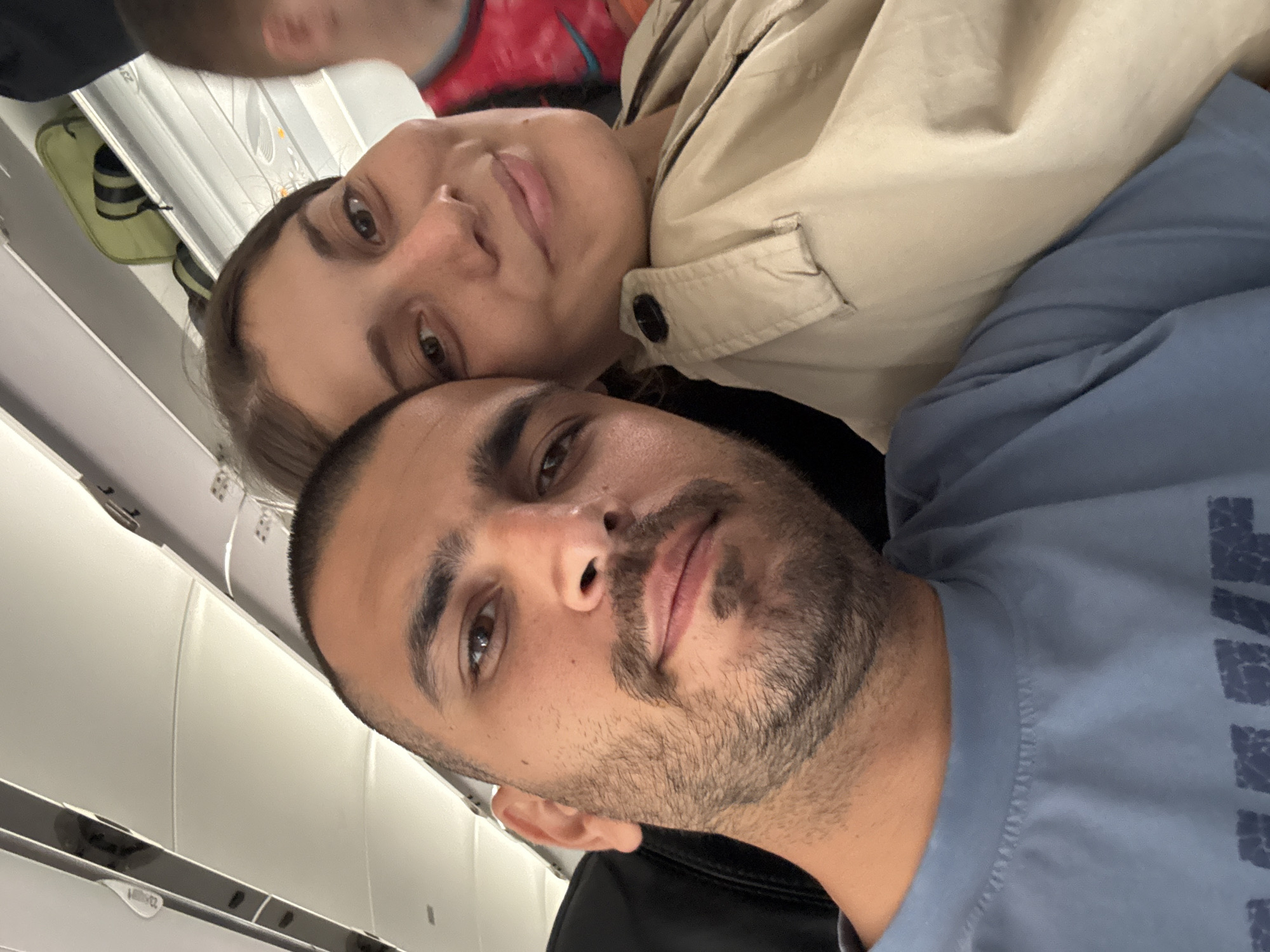 A selfie depicts a couple aboard an airplane. The man, with dark hair and a beard, is wearing a blue t-shirt. Beside him, a woman with fair skin and brown hair is wearing a beige jacket. The interior of the airplane cabin is visible in the background, indicating the location of the subjects. The location is not specifically identifiable as Đà Nẵng, Vietnam from this image alone.