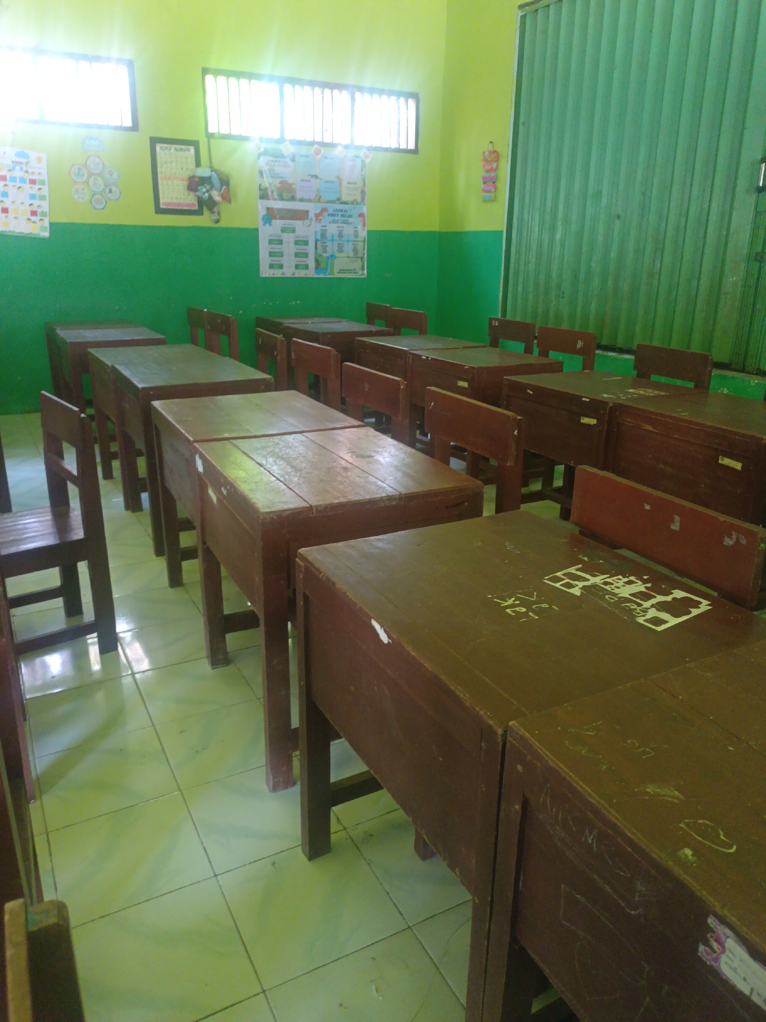A classroom in Sarimulyo, Indonesia, is depicted, featuring rows of empty wooden desks and chairs. The walls are painted lime green above a green wainscot. Posters are affixed to the walls, including a colorful chart with figures and a framed schedule. Sunlight streams through barred windows, casting a bright glow. A tall, green slatted door or window covering dominates the right side of the frame. The floor is tiled in a pale green hue. The overall impression is one of a simple, functional learning environment.