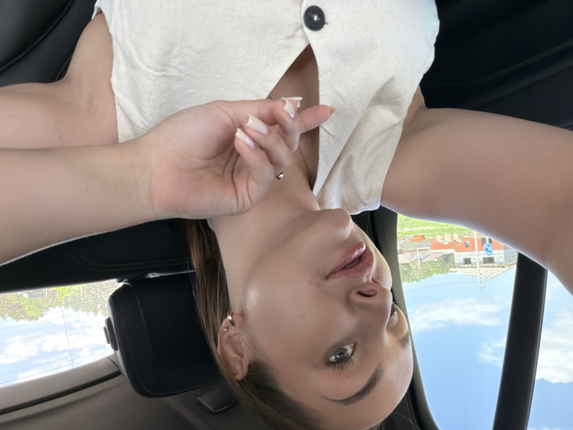 A woman is taking a selfie in the back seat of a car. She's wearing a cream-colored shirt with a button. The image appears to be taken at an angle and upside down. Outside the car window, one can see buildings and the sky. The geographical location is likely Chaikovsky, Russia.