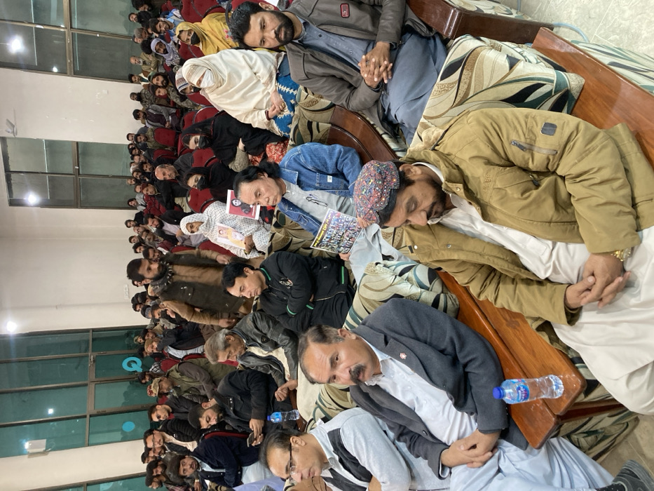 The image depicts an indoor gathering of approximately 60 to 70 individuals, predominantly adult males, within a large hall in Quetta City Tehsil, Pakistan. Attendees are seated in rows on upholstered benches with wooden frames and some individual chairs, generally oriented towards an unseen focal point.
In the foreground, several men are seated. A man on the far right wears a brown jacket over a light-colored shalwar kameez and a colorful, embroidered cap. His hands are clasped. Next to him, a man with a mustache wears a grey suit jacket over a light-colored shalwar kameez. A plastic water bottle (Nestlé Pure Life brand) rests on the wooden armrest between them. To the left, a man in a grey long-sleeved shirt and glasses is visible.
In the mid-ground, a woman wearing a white head covering (dupatta or shawl) is seated, holding a portrait-style photograph of an individual. To her right, another individual holds a rectangular sheet displaying multiple small photographs arranged in a grid. Other individuals in the mid-ground include men in denim jackets, dark tracksuits, and various traditional and casual attire. Some women are also visible, wearing dark headscarves or light-colored shawls.
The background shows additional rows of seated attendees, densely filling the hall. The rear wall features large, light-colored panels, some of which appear to be tall glass windows or reflective surfaces, exhibiting internal reflections. The ceiling is light-colored and high, with visible artificial lighting fixtures. The overall environment is well-lit.