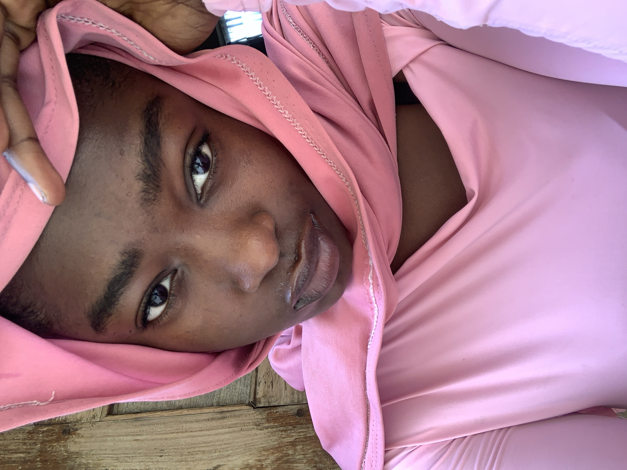 A close-up, tilted portrait shows a Black woman wearing a pink hooded garment. The wooden surface beneath her suggests an outdoor setting. The location, Rafin Par, Nigeria, is not discernible from the image.