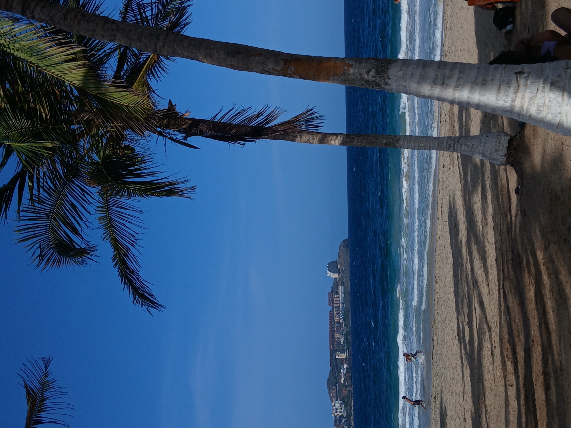 This image captures a sunny day at a beach in Pampatar, Venezuela, with the Caribbean Sea in the background. The foreground is dominated by the trunks of two palm trees, casting shadows on the sand. In the middle ground, the turquoise ocean meets the sandy shore with gentle waves. Two figures, appearing to be young males, are visible in the water near the shore, one running into the sea and the other splashing. In the distance, across the water, a cityscape is nestled on a hill, characterized by various buildings and structures, suggesting a developed coastal town. The sky is a clear, vibrant blue, indicating good weather.