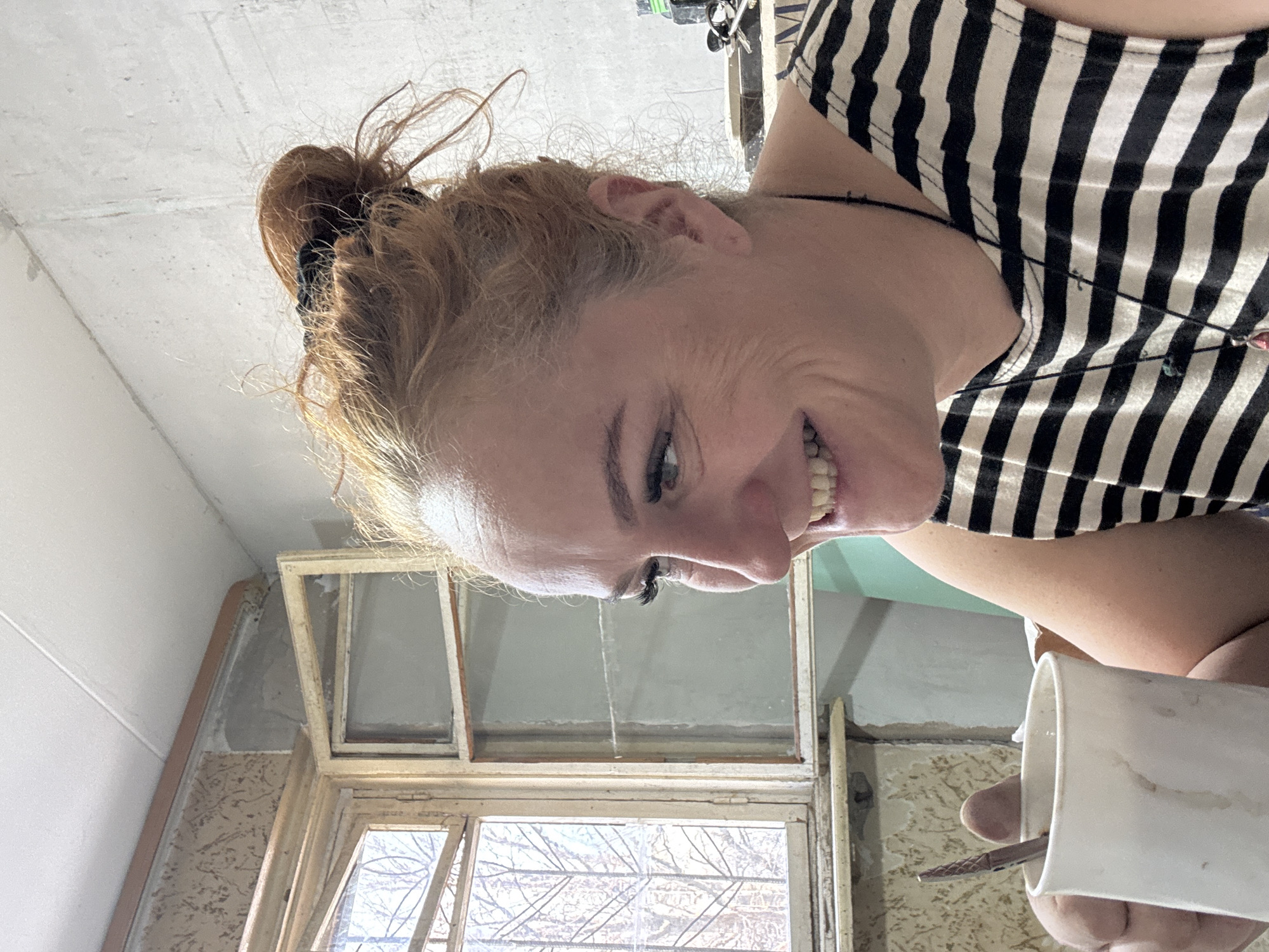 A woman with red hair in a bun, wearing a black and white striped shirt, is holding a white mug with a tool inside, and is smiling. She is positioned near a window with bare tree branches visible outside. The surrounding walls show signs of renovation or construction, possibly a residential interior. The specific location within the city of Chaikovsky, Russia, cannot be determined from the image.