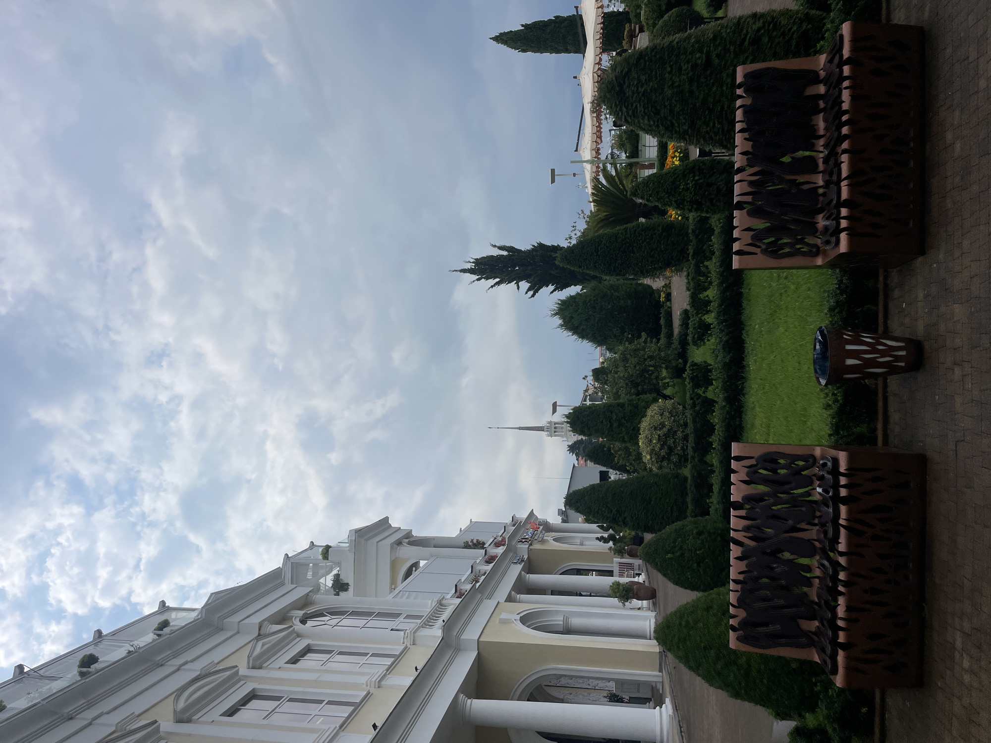 The image depicts an exterior view of a building in Sochi, Russia, likely the former Soviet health resort building, now part of the Radisson Collection Paradise Resort & Spa, Sochi. The building is grand, with a white facade, arched windows, and classical columns lining the lower level. The sky is overcast with soft, scattered clouds. In the foreground, a manicured garden features lush green lawns, neatly trimmed bushes, and several topiary trees. Two unique, modern benches with intricate, cut-out designs in a metallic brown color are positioned facing the lawn. Between the benches, a patterned trash receptacle is visible. In the midground, a spired building, possibly a church or a landmark, rises above the trees. The overall atmosphere is serene and well-maintained.