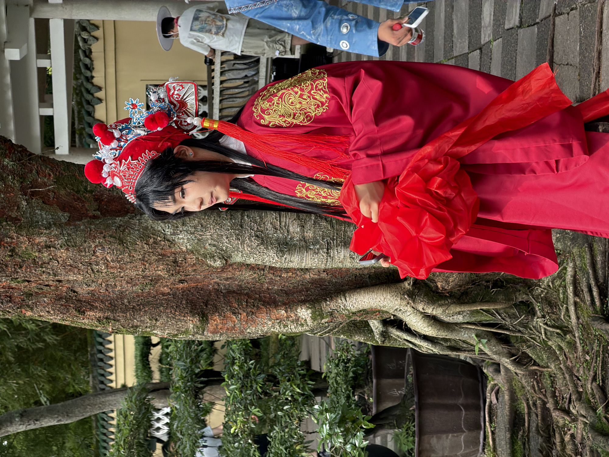 In Yuzhong District, China, a young person dressed in traditional red opera attire, complete with an elaborate headdress, stands near a large, gnarled tree. The individual holds a crumpled red fabric in their hands and appears to be looking to the right. Behind them, a person in a white t-shirt and shorts with a hat is visible, and another individual in a blue garment is also present, holding a beverage can and a phone. The background features lush greenery, potted plants, and architectural elements of what appears to be a garden or cultural site, with a tiled roof and pillars. The ground is paved with stone.