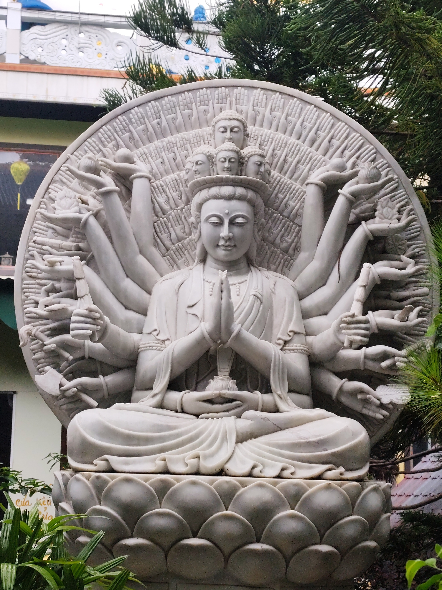 The central focus of the image is a large, intricately carved white marble statue of a multi-armed Buddhist deity, most likely Guanyin (Avalokiteshvara). The deity is depicted in a serene, meditative pose, seated cross-legged on a prominent lotus pedestal, with the primary hands joined in a prayer mudra.
Numerous additional arms extend from the sides and back of the main figure. Many of these hands hold symbolic objects, including a lotus flower, an axe, an orb, a staff, and a feather. Behind the main figure, a large, circular halo-like structure is filled with hundreds of smaller, finely detailed hands, each seemingly holding an eye in its palm, symbolizing wisdom and universal compassion. Several smaller heads are carved above the main deity's head, stacked vertically.
The statue is set outdoors, surrounded by vibrant green foliage, including dense pine-like trees that frame the top and sides. In the background, parts of a building with ornate white architectural elements and blue accents are visible, suggesting a temple or pagoda structure. A distinct yellow lantern hangs from a distant building on the left. The scene is illuminated by diffused daylight, indicating an overcast sky or soft lighting conditions. Faint Vietnamese text, possibly "Cửa Kẽm ?", can be seen on a white surface in the lower-left corner. The overall impression is one of peaceful devotion and detailed craftsmanship, situated within a natural and spiritual environment typical of a temple in Da Lat, Vietnam.