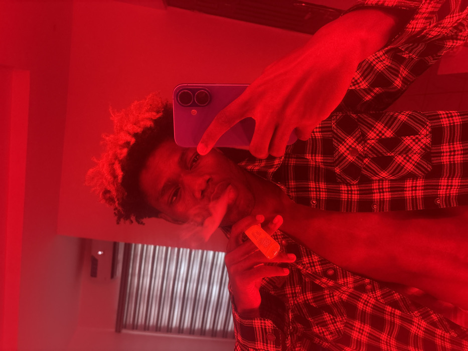 This portrait-oriented image, heavily saturated with a red filter, depicts a single male individual indoors in Igboefon, Nigeria, appearing to take a mirror selfie. The subject, a dark-skinned male with styled hair featuring lighter tips, is centered in the frame. He wears an unbuttoned red and black plaid long-sleeved shirt, exposing his chest. In his left hand (viewer's right), he holds an electronic vaping device, with vapor emanating from his mouth. His right hand (viewer's left) holds a light-colored smartphone, possibly purple, displaying two prominent rear camera lenses, and forms a "V" or "peace" sign gesture. The background consists of an indoor room with walls illuminated uniformly by a strong red light. To the upper left, a white air conditioning unit is mounted on a wall, and below it, a window with horizontal blinds is partially visible.