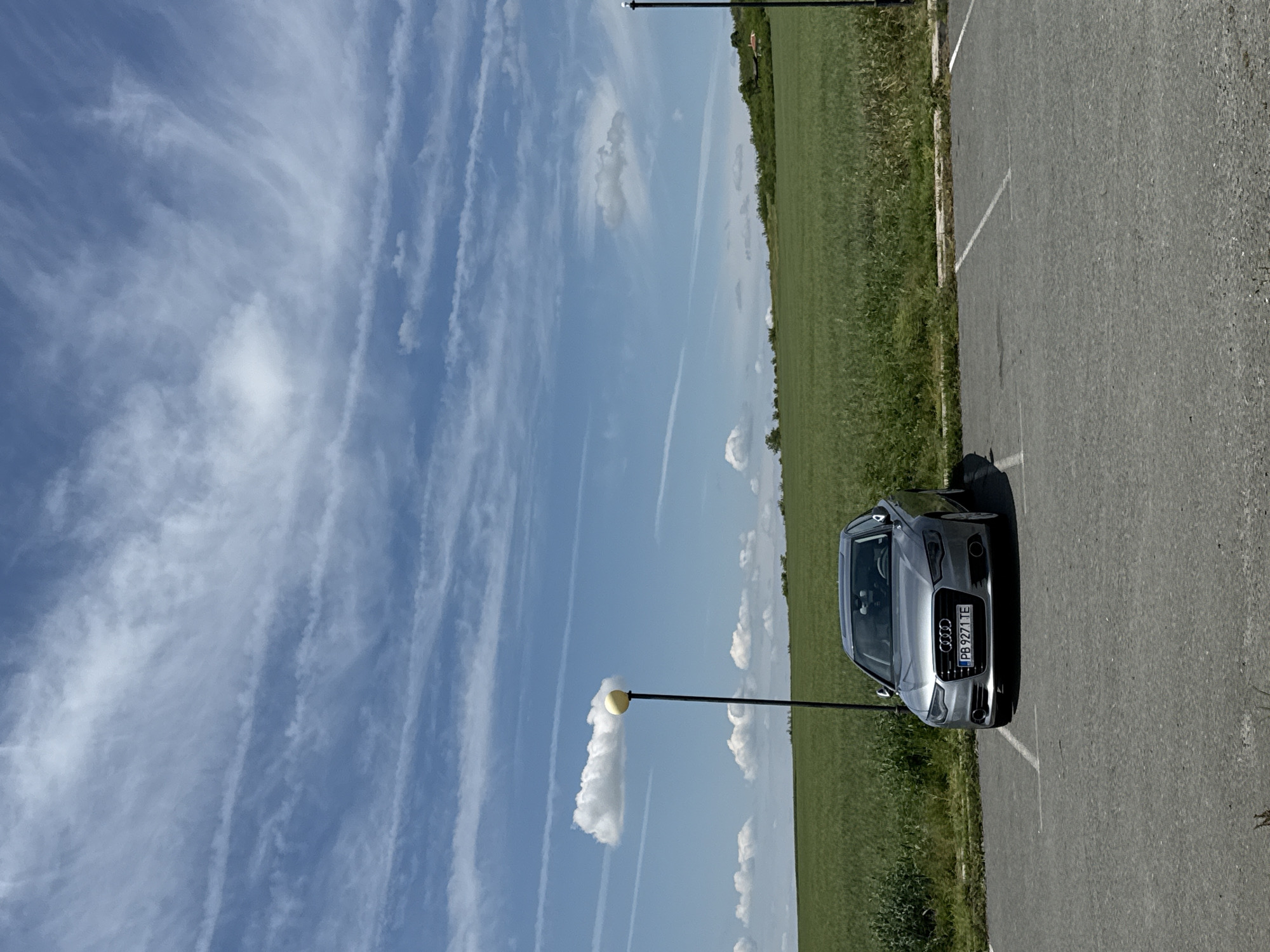 A daytime shot of a silver Audi parked in a parking space. The vehicle's license plate reads "PB 9271 TE". A mowed green field borders the parking area, next to a blue sky with striated clouds. A single yellow light post stands to the left of the car. Based on the license plate and the general environment, the location could be Kabile, Bulgaria, but that is not verifiable from this image alone.