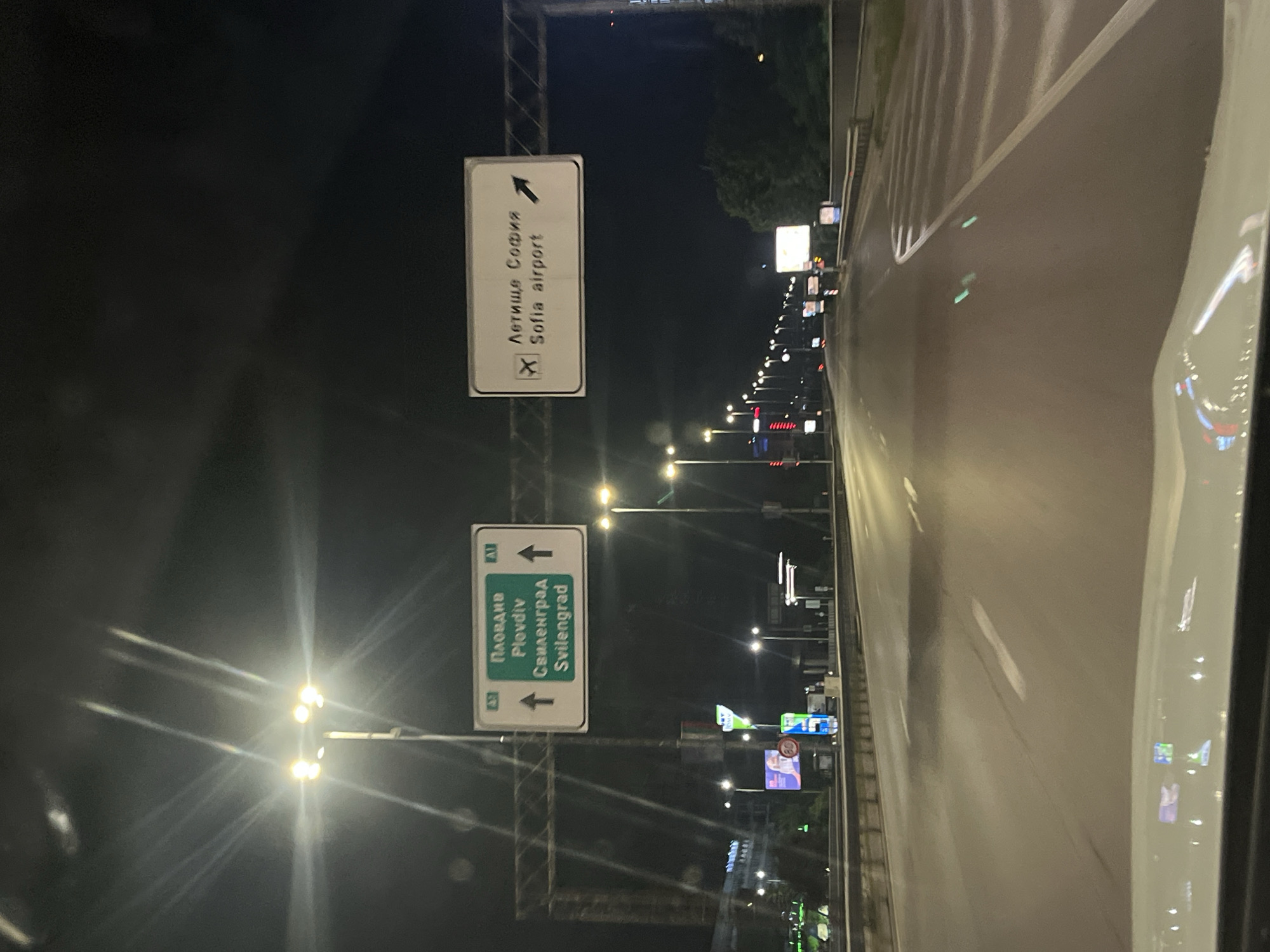 The image shows a road at night in Sofia, Bulgaria. Two signs are visible above the road. The top sign indicates the direction to Sofia Airport. The bottom sign indicates the direction to Plovdiv and Svilengrad. Streetlights line the road. The picture was likely taken from inside a vehicle.