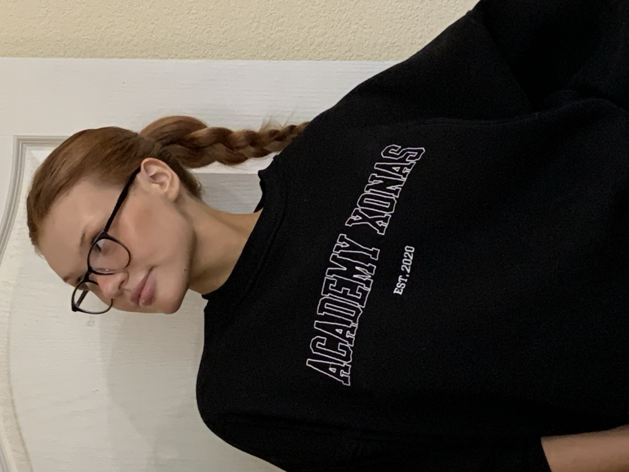 The image depicts a young female indoors in what is presented as the city of Chaikovsky, Russia. She has reddish-brown hair styled into a single, thick braid positioned on her right side (viewer's left). Wearing dark-framed eyeglasses, she looks upward and to her left, with her lips slightly parted. Her attire consists of a black, long-sleeved sweatshirt. Prominently displayed across the chest of the sweatshirt are the words 'ACADEMY XONAS' in large, white, outlined capital letters, angled slightly upward from left to right. Below this text, in smaller, solid white lettering, is 'EST. 2020'. The background shows an interior setting with two distinct wall textures: a white wall with subtle detailing and a decorative arch or molding on the left, and a coarser, textured beige wall on the right. These wall sections are separated by a vertical white door frame. The lighting suggests an artificial indoor source.