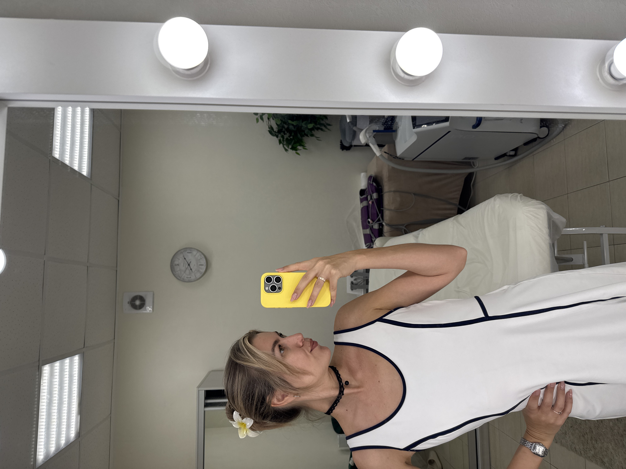 The photograph captures a woman taking a selfie in front of a vanity mirror. The location appears to be an interior setting, possibly a beauty salon or dressing room in Unknown, Unknown. The mirror is framed with illuminated light bulbs across the top. Reflected in the mirror is the woman holding a yellow smartphone. She is wearing a white dress with black trim, a beaded choker necklace, and a watch. Her hair is styled with a white flower. In the background, there is a wall with a clock, an air conditioning vent, and a fluorescent light panel. A medical apparatus and a covered bed are visible further in the background. The overall scene suggests a clinical or cosmetic setting.