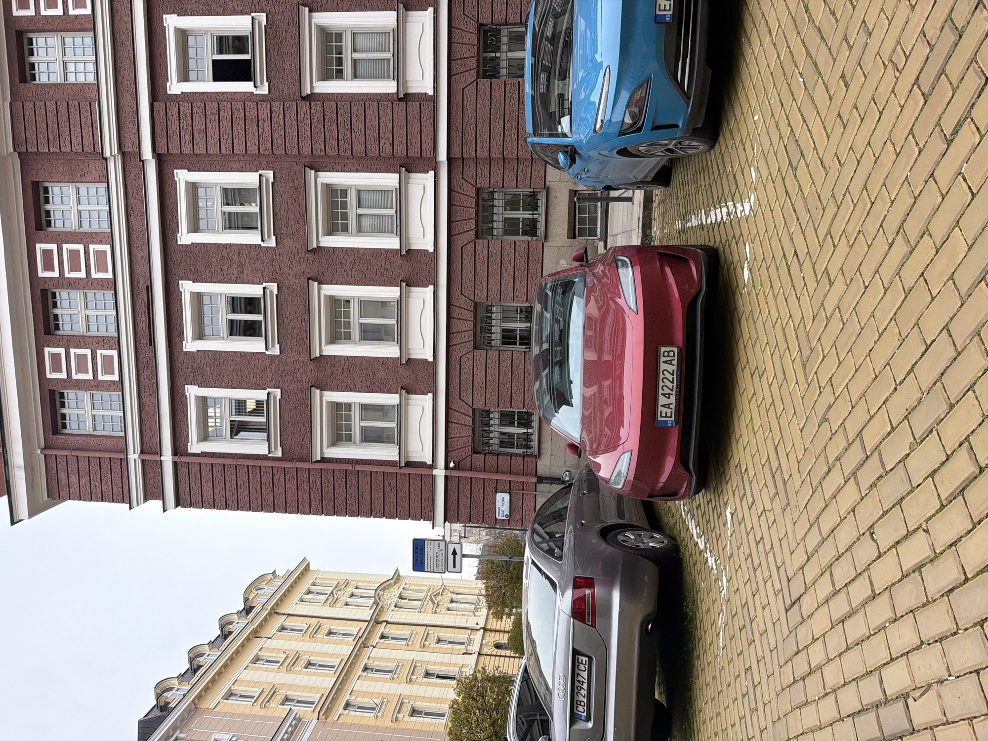 The image shows a street in Sofia, Bulgaria. Three cars are parked along a yellow brick road, in front of a building with red brick walls and white window frames. The cars are a red Tesla, a blue Hyundai Kona and a gray Audi. A street sign is visible between the red and gray cars, indicating a "BLUE ZONE". Another building with light yellow walls and white windows is visible across the street. The sky is overcast.