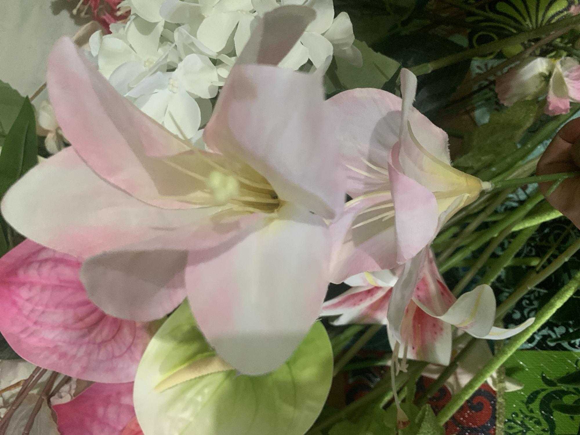 A close-up, overhead shot displays a floral arrangement dominated by large, pale pink lily-like flowers. These flowers feature prominent yellow stamens and delicate pink veining on their petals. To the left, a bright green anthurium bloom with a yellow spadix is partially visible, along with a pink flamingo anthurium. In the background, clusters of small, white flowers, possibly hydrangeas, add texture and contrast. The stems of the flowers are a vibrant green, and some are held by a person's hand at the bottom right of the frame. The arrangement is set against a background with hints of green and patterned fabric, suggesting an indoor setting. The overall impression is of a lush, vibrant, and somewhat artificial floral display.