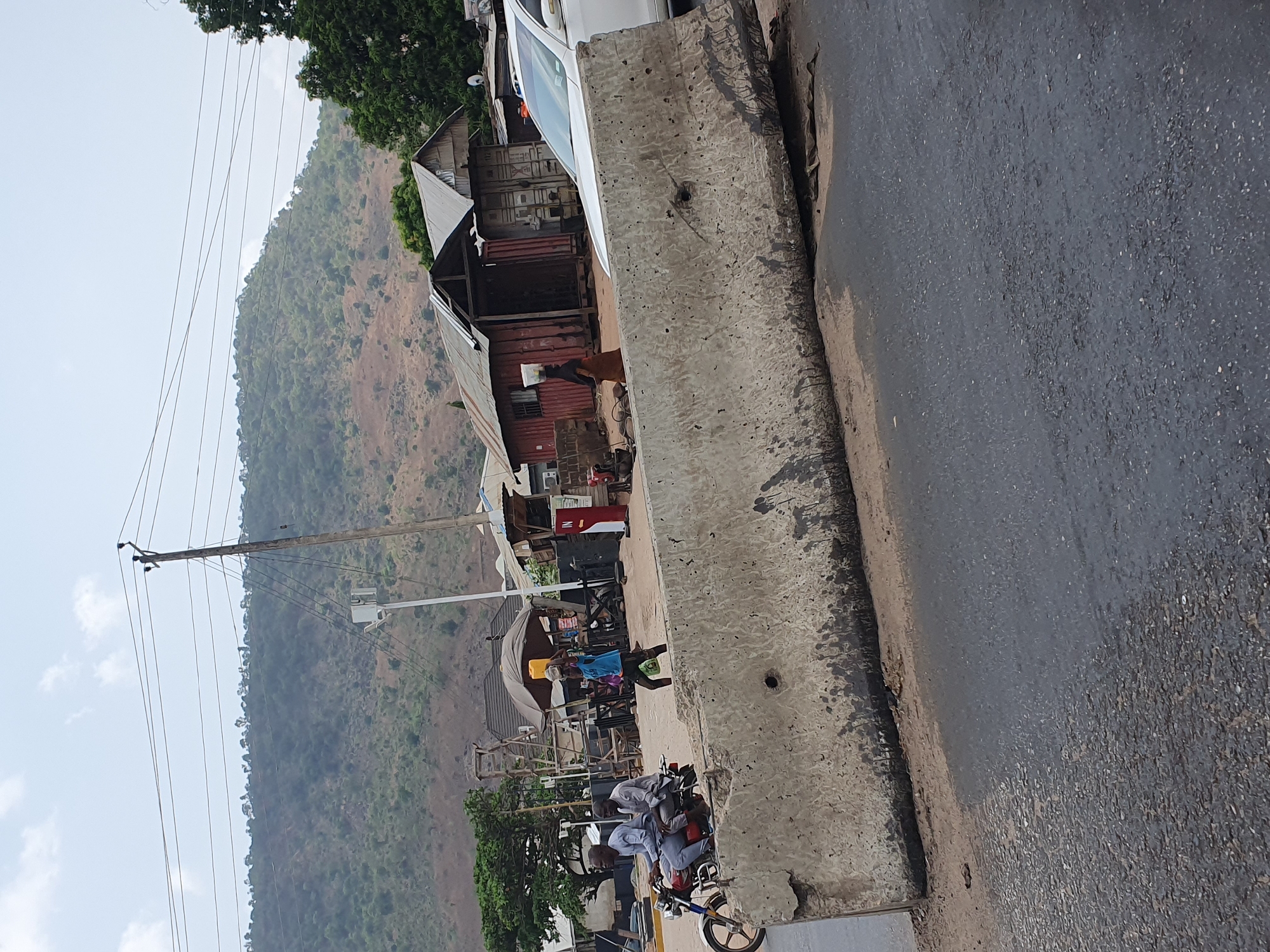 The image depicts a street scene in Lokoja, Nigeria. A concrete barrier divides the foreground from the street and some buildings. The road surface is dark and textured, showing signs of wear. Behind the barrier, there is a mix of informal commercial establishments, including structures made from shipping containers and corrugated metal. People are present, some on motorcycles. A mountain covered in green vegetation is visible in the background. Overhead power lines run across the scene.