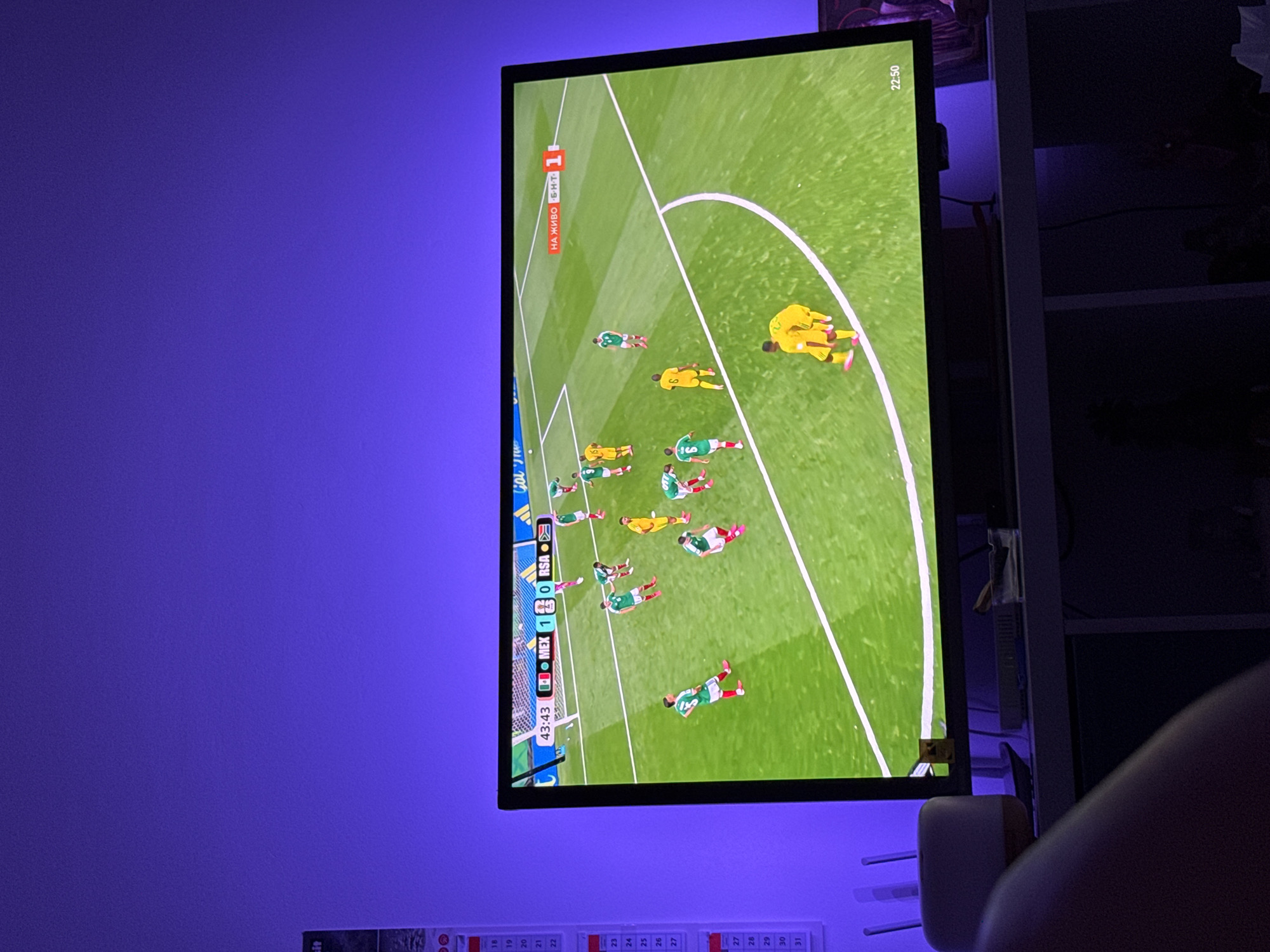 In the city of Plovdiv, Bulgaria, a television displays a football match. The screen shows a game between Mexico (MEX) and South Africa (RSA), with the score at 1-0 in favor of Mexico, and the time at 43:43. Several players in green and white kits, representing Mexico, are on the field. Two players in yellow kits, representing South Africa, are also visible. The match is being broadcast live, indicated by the "НА ЖИВО" (live) text on the screen. The television is placed on a shelf in a dimly lit room, with ambient purple lighting emanating from behind the screen. The background suggests a domestic setting, with a calendar visible on the left wall and a router and a small item on the shelf below the television.