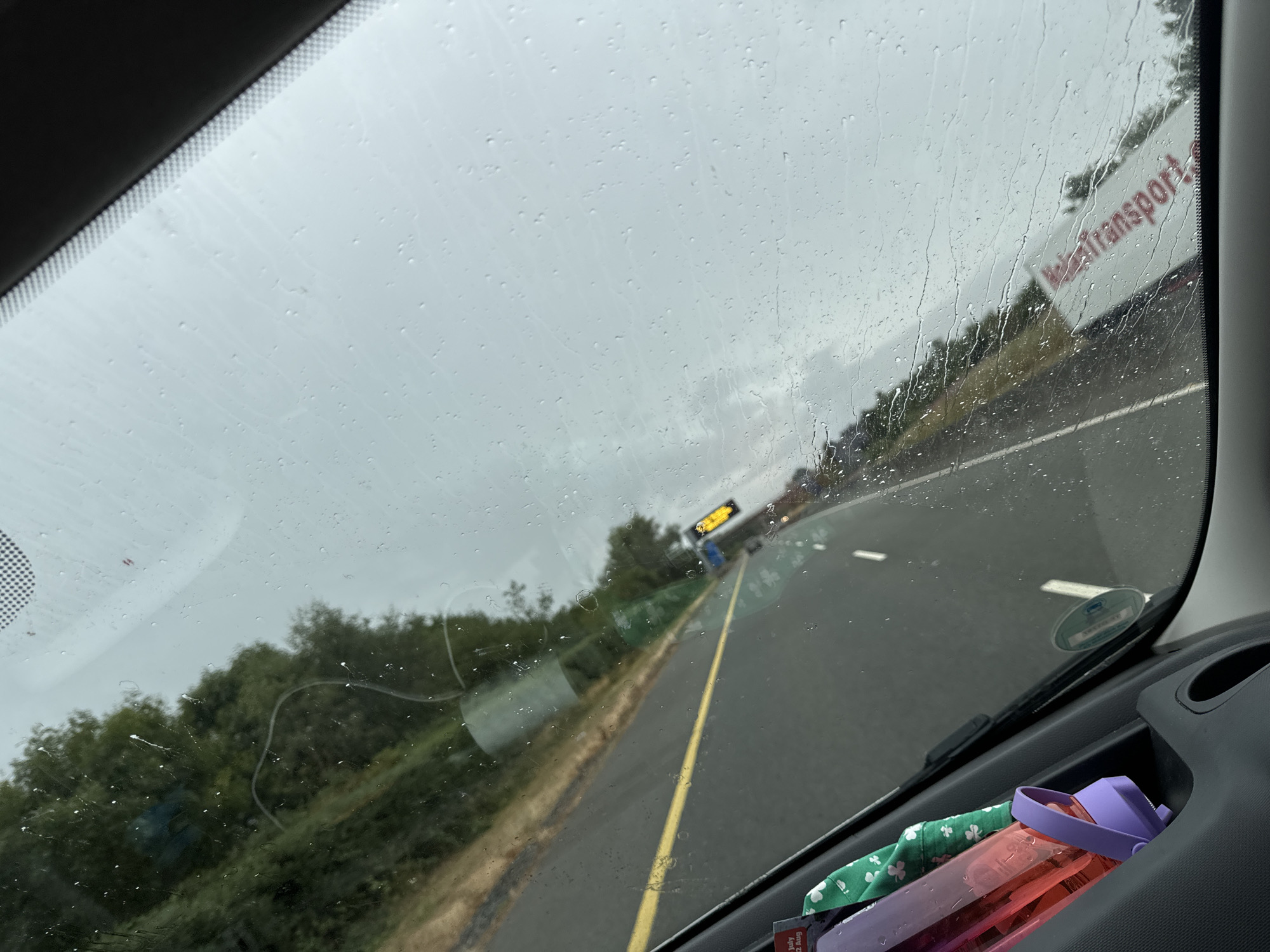 The image shows a view from inside a vehicle, looking through a rain-streaked windshield. The road ahead is visible, with a yellow line marking the lane. Trees and foliage line the side of the road. In the distance, there is a traffic jam with a truck that has "Transport" written on its side. A yellow digital sign is also visible on the right side of the road. Inside the vehicle, a purple and pink water bottle and a green cloth with a shamrock pattern are visible in the lower left corner. The sky appears overcast.