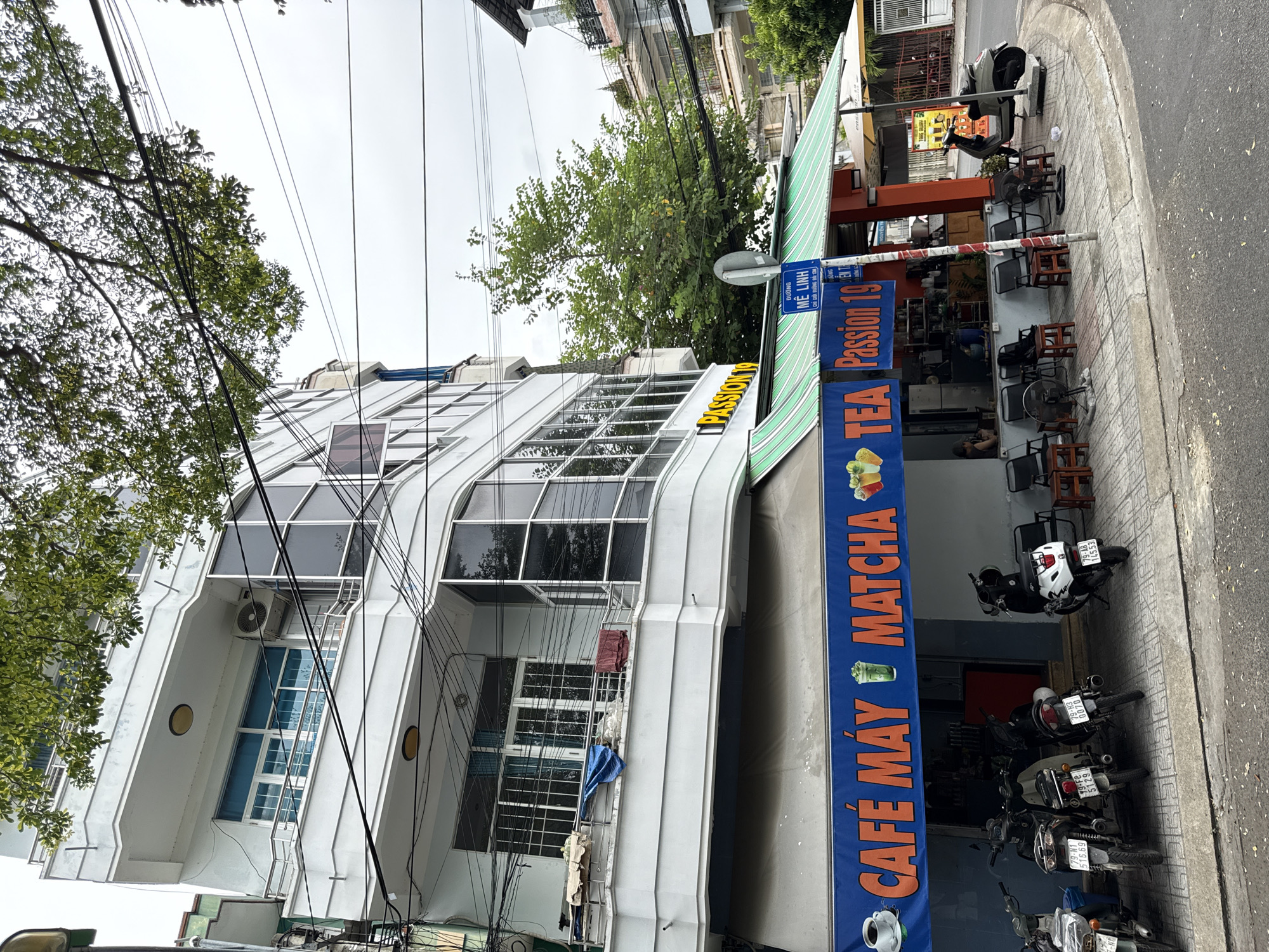 The image shows a street scene in Nha Trang, Vietnam, featuring a multi-story building with a cafe on the ground floor. The cafe, identified by a blue banner with orange lettering that reads "CAFÉ MÁY MATCHA Passion 19", has an outdoor seating area. Several motorbikes are parked along the sidewalk in front of the cafe. A green and white striped awning extends over the seating area. A blue street sign with Vietnamese text is visible on a pole. Trees and overhead wires are present, indicating an urban environment. The sky is overcast.