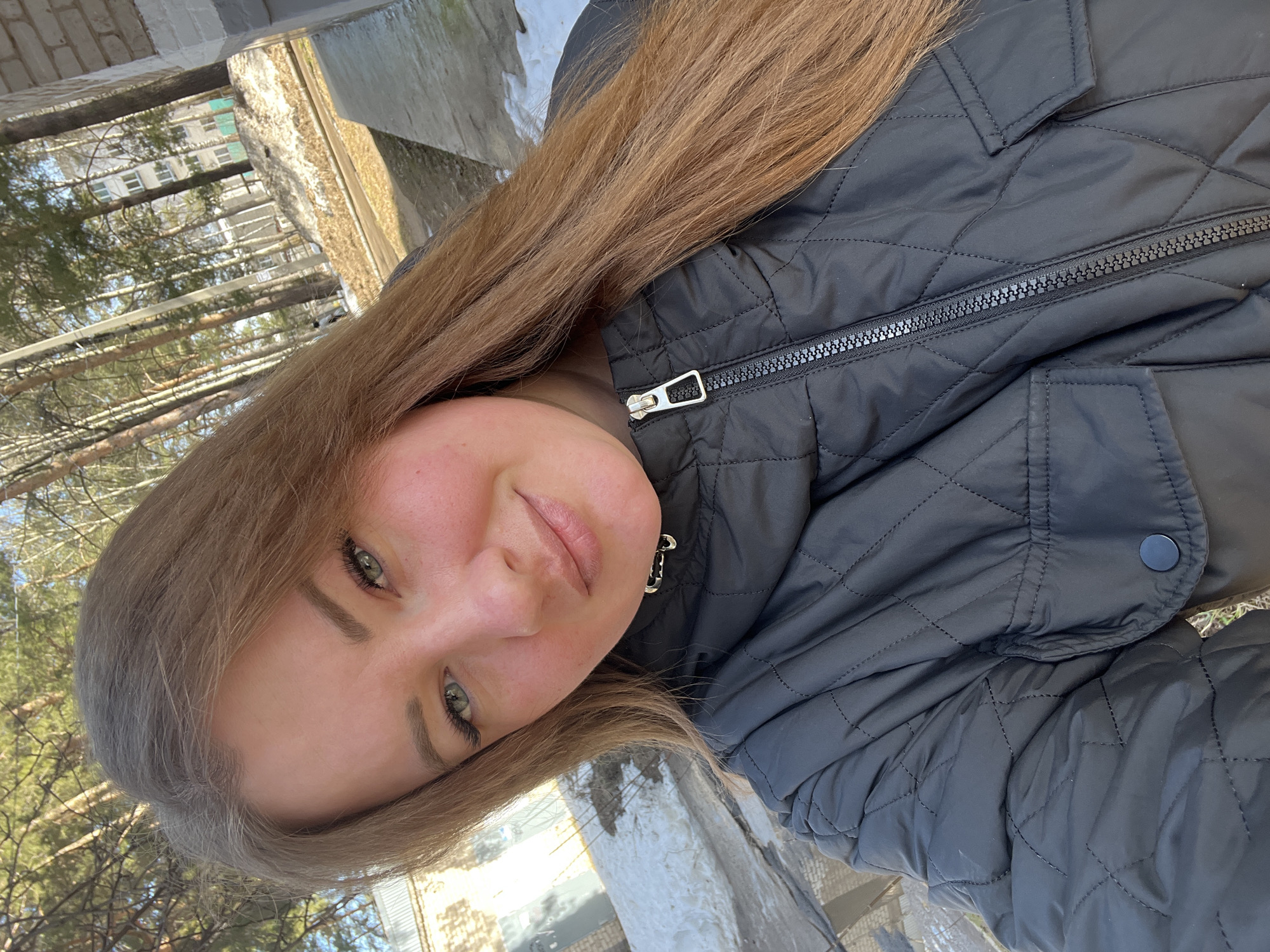 The image shows a woman with long, light brown hair, fair skin, and green eyes. She is wearing a black quilted jacket with a zipper and button detail. The background includes trees, a building, and what appears to be a concrete structure, potentially a wall or ledge. Given the location of Chaikovsky, Russia, the presence of trees and the type of building suggest a residential area or a park-like setting within the city. The weather seems to be bright with some snow in the background. The image is taken at an angle, suggesting a selfie or a picture taken by holding the camera at a tilted position.