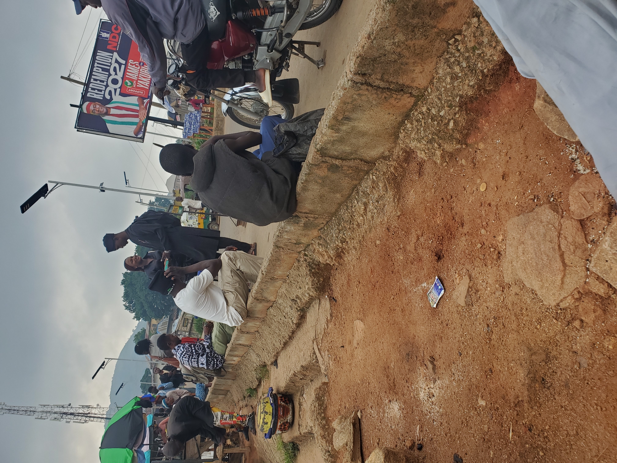 In Gwong, Nigeria, an outdoor scene unfolds with a group of people gathered on a street. A billboard prominently displays "REDEMPTION 2027" with an image of "HON. JAMES YAKUBU" and the NDC logo. Several individuals are seated on a concrete curb, facing the street. Among them, a man in a black hooded vest and blue pants faces away from the camera, while another man in a white t-shirt and khaki pants is also seated. A motorcycle with a rider is parked nearby, partially obscuring another person. In the foreground, the ground is covered in reddish-brown dirt and rocks, with some scattered debris, including a colorful plastic bowl and what appears to be a discarded packet. In the background, a large telecommunications tower is visible, along with market stalls and buildings, suggesting a bustling urban or semi-urban environment. The sky is overcast, indicating a cloudy day.