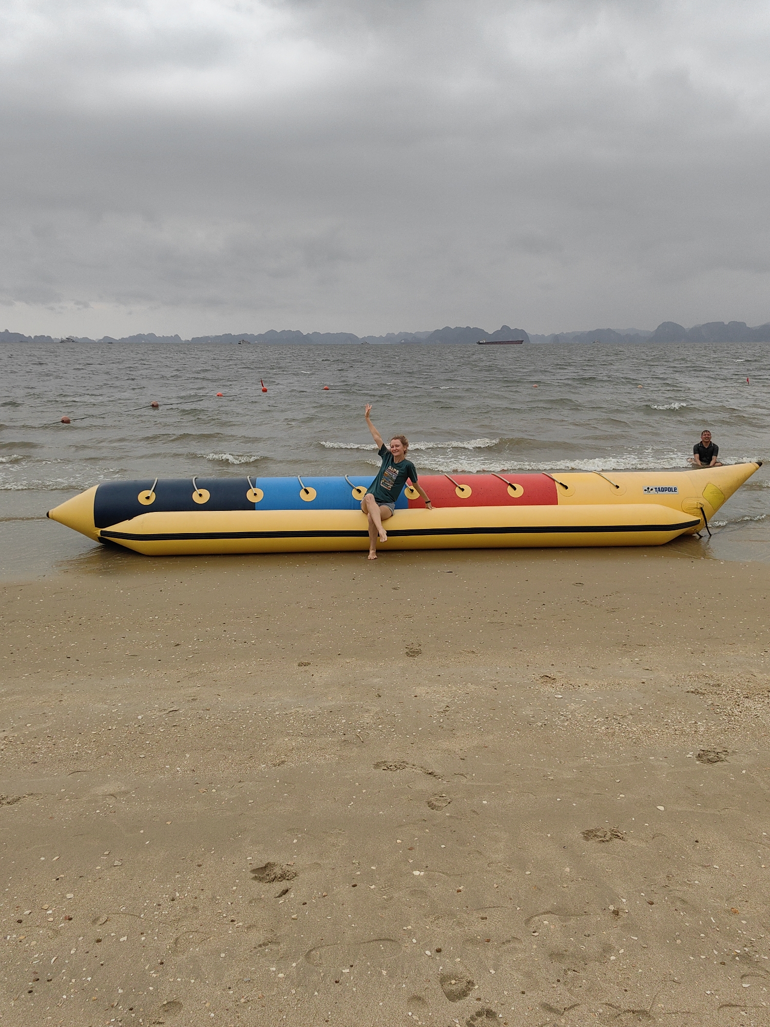 A beach in Bai Chay Ward, Vietnam on an overcast day shows a large inflatable banana boat resting on the sand with the bow partially in the water. A woman in a dark shirt and shorts sits on the banana boat, waving. Further towards the tip of the banana boat in the water sits another person. The banana boat is yellow with black, blue, and red sections on top, and the word "TADPOLE" is printed on the side. The beach is tan sand with small shells and pebbles. In the background, the sea is choppy and islands are visible. A cargo ship is visible on the horizon. Orange buoys are floating in the water.