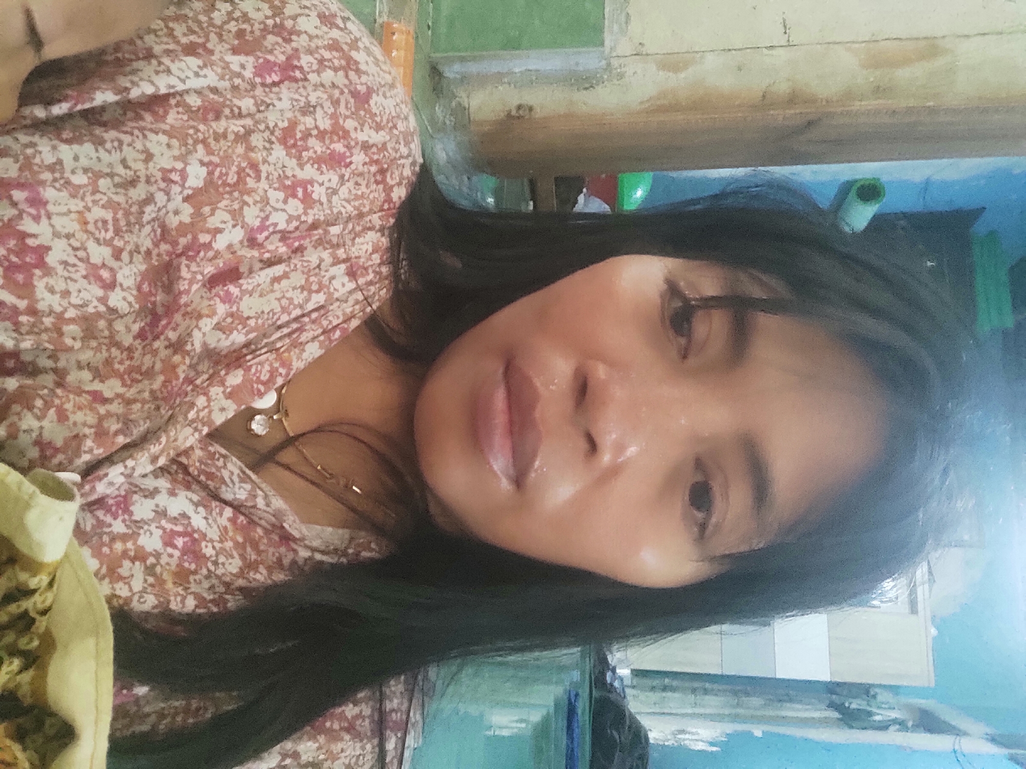 The image is a close-up, selfie-style photograph of a woman with dark hair and a floral print shirt. She is wearing a gold necklace with a pendant. The background appears to be an indoor environment with a blue wall and some wooden elements. The lighting suggests it might be daytime, with some harsh light creating reflections on her skin. The overall impression is that of a casual, personal photograph taken in what could be a residential setting in Sakra, Indonesia.