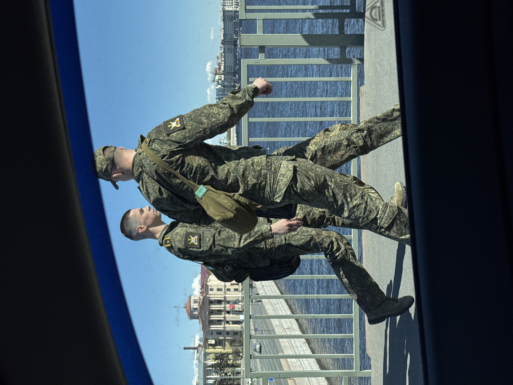 Two men in camouflage military uniforms stand on a bridge with a metal railing overlooking a body of water. The skyline of Saint Petersburg, Russia, is visible in the background, including structures such as the Nakhimov Naval School. The men wear military caps, and one carries a shoulder bag. They appear to be looking out at the water and the city. The image is taken from inside a vehicle, as parts of the car frame are visible on either side.