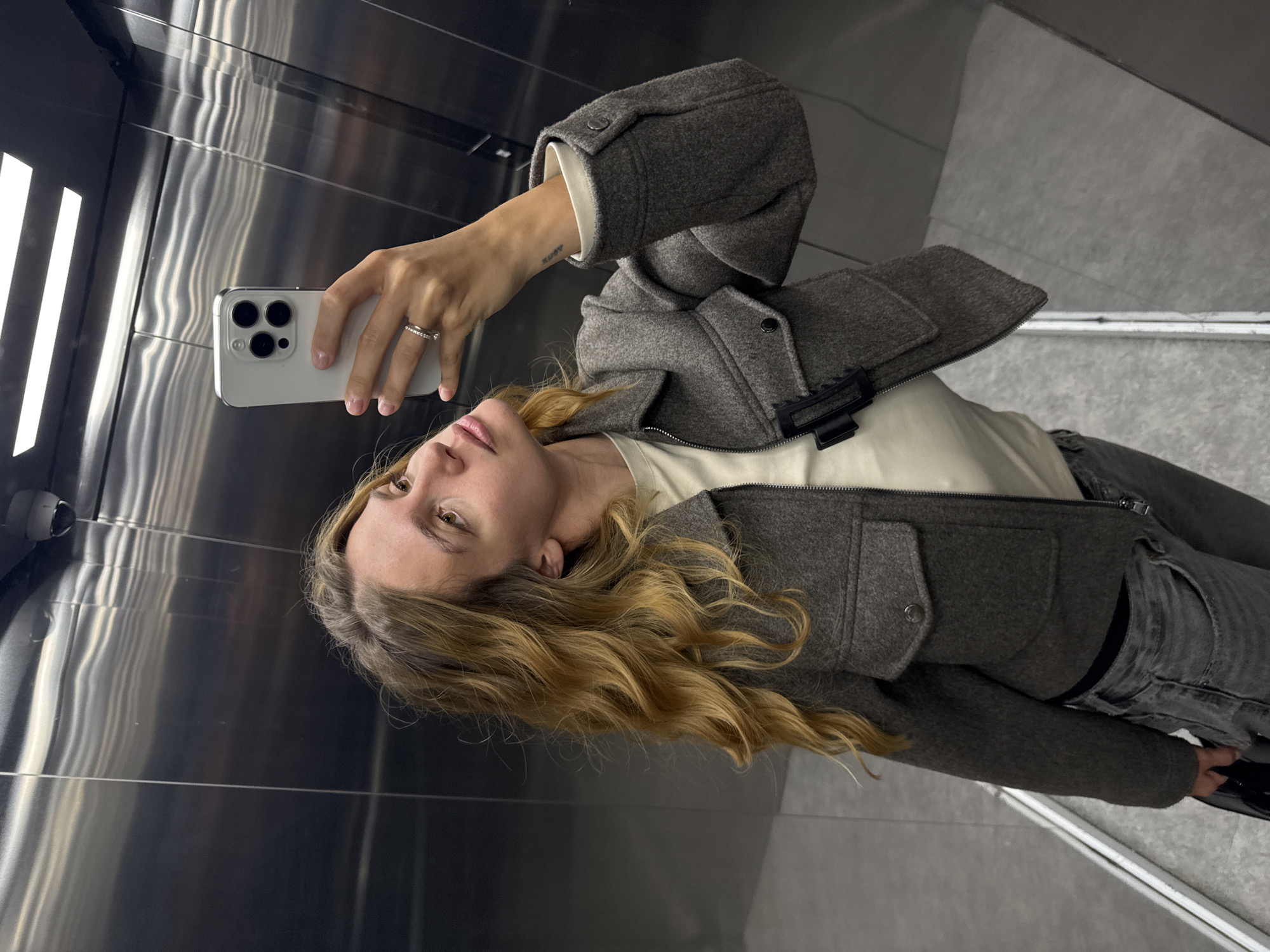 A woman is taking a selfie in an elevator. She is holding a white smartphone in her right hand. She is wearing a gray jacket, a white shirt, and gray jeans. The elevator has shiny metallic walls and a white light panel. There is a security camera in the upper left corner. The location is Unknown, Unknown.