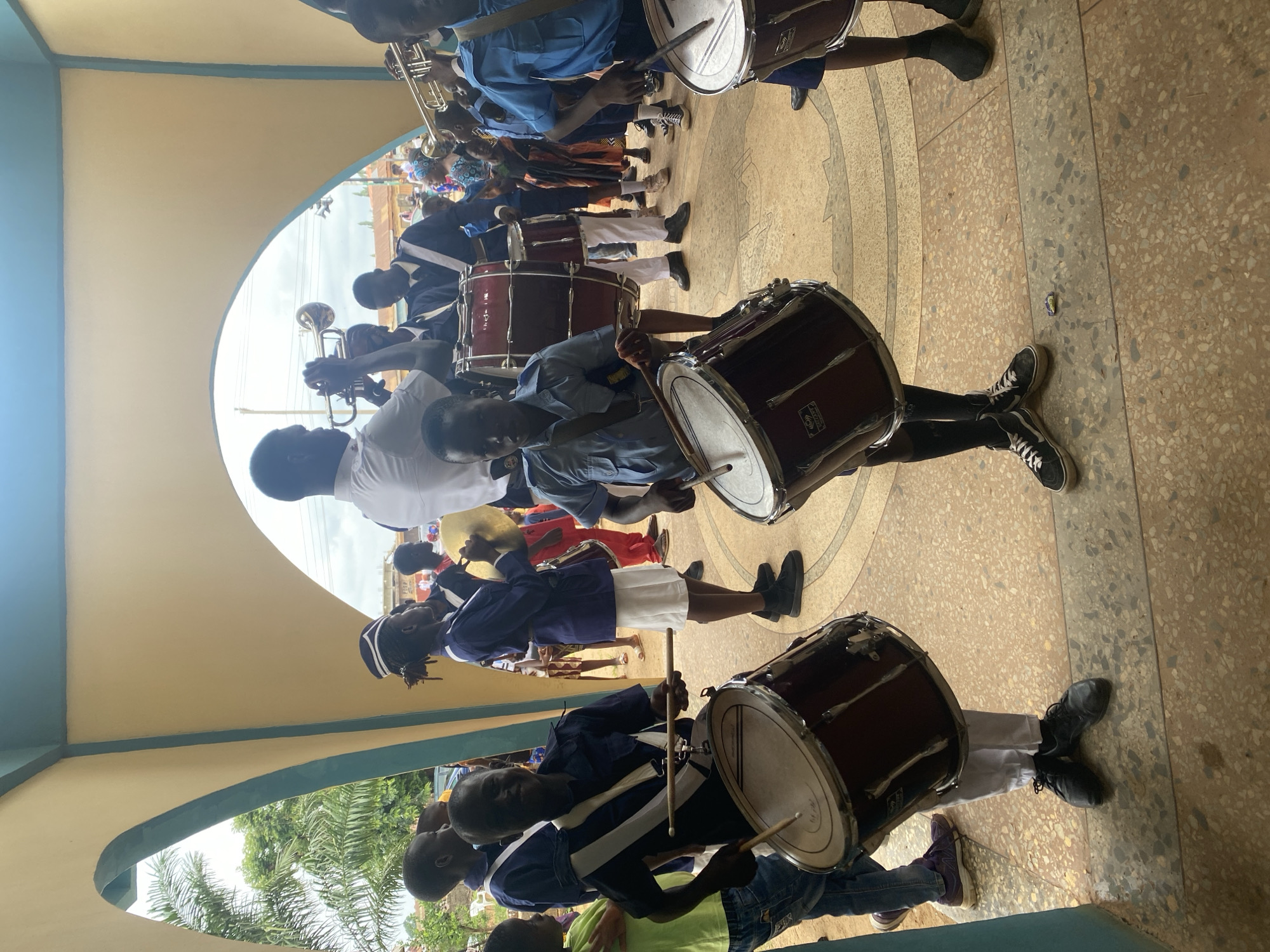 A marching band is assembled under an archway in what appears to be Kafanchan B, Nigeria. The band consists of young people, primarily dark-skinned, playing drums and trumpets. Some members are wearing white, blue, and purple uniforms with black shoes. They are positioned on a tiled floor with circular patterns. The archway has a yellow-painted ceiling with blue trim, and palm trees are visible in the background through the arch.