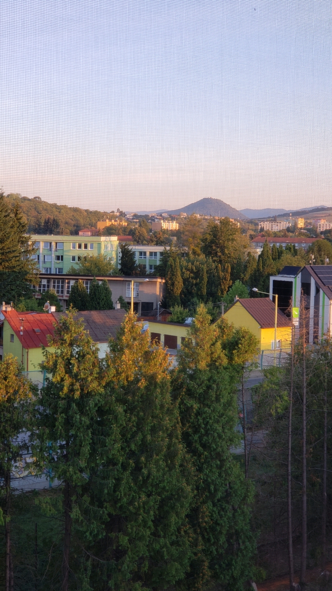 This image captures a view of a residential area in Prešov, Slovakia, likely taken from an elevated position like a window, as indicated by the mesh pattern superimposed on the scene. The time of day appears to be late afternoon or early evening, judging by the warm sunlight casting long shadows and illuminating the foliage.
In the foreground, dense evergreen trees dominate, their branches a vibrant green, catching the golden light. Behind them, a mix of residential buildings can be seen. To the left, a low-slung building with a bright red roof and yellow siding is visible. Adjacent to it is a larger building with a brown roof and light-colored facade. Further back and slightly to the right, a prominent bright yellow building with a brown roof stands out.
Behind these closer structures, several larger apartment buildings are present. Two prominent buildings with green facades and multiple windows are central in the mid-ground. Other buildings, some with lighter colors and varying architectural styles, are interspersed among the trees. The background features a hilly landscape with a distinct conical hill dominating the skyline in the distance. The sky is a clear, pale blue, transitioning to a lighter hue near the horizon. The overall impression is a peaceful suburban or semi-urban setting within Prešov.