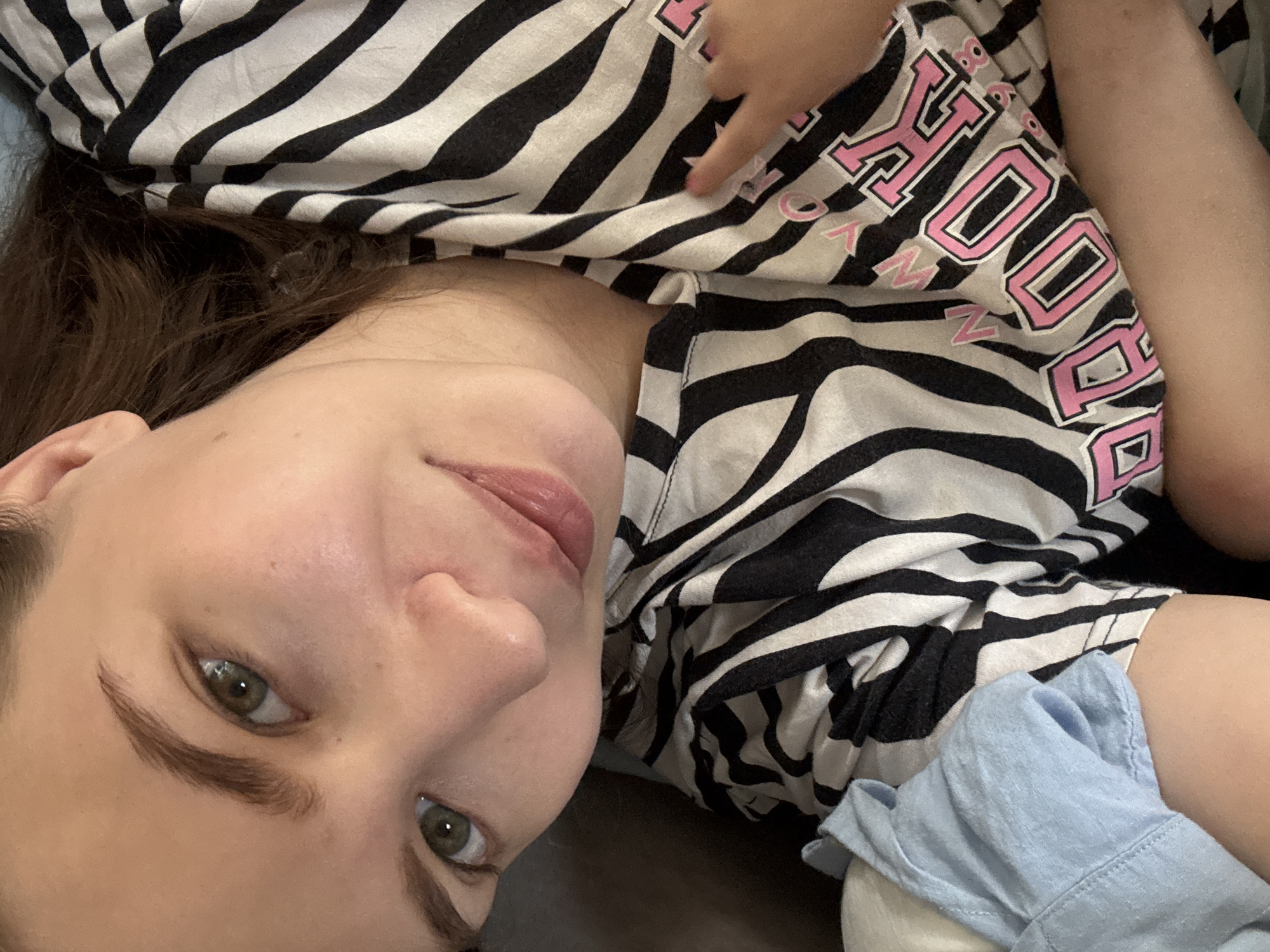 A close-up, high-angle selfie shows a person lying down, their face taking up most of the frame. They have light skin, green eyes, and brown eyebrows. Their hair is dark brown and falls around their face. They are wearing a black and white striped shirt with pink lettering that appears to spell out "BROOKLYN." A blue garment is visible on the left side of the frame, partially obscuring the person's arm. The background is dark, suggesting an indoor setting. There is no identifiable information about the location in the city of Chaikovsky, Russia.