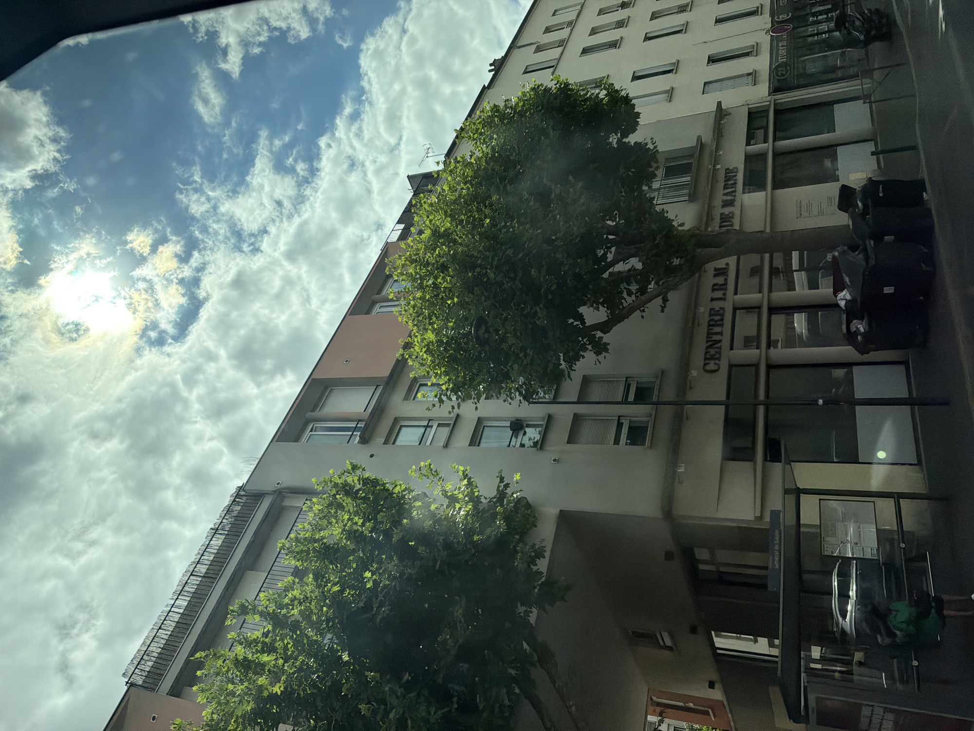The image shows a building with trees in front of it. The building appears to be an apartment complex or office building with multiple floors and windows. There is a street in front of the building, and some luggage or bags are placed on the sidewalk. The sky is visible in the background, with clouds and the sun shining through.
The building has a sign that reads "CENTRE I.R.M. DE MARNE". The architecture suggests an urban or suburban setting. The presence of luggage might indicate travel or moving. The trees are leafy and green, suggesting it is daytime and likely spring or summer. The sky appears bright, indicating it's daytime, possibly late morning or early afternoon given the sun's position.
The scene is captured from a low angle, looking up at the building and the sky. The overall mood is neutral, presenting a typical street view of a building in Alfortville, France. No people are clearly visible, though the presence of luggage suggests activity nearby. The image is in color and appears to be taken with a camera or smartphone.