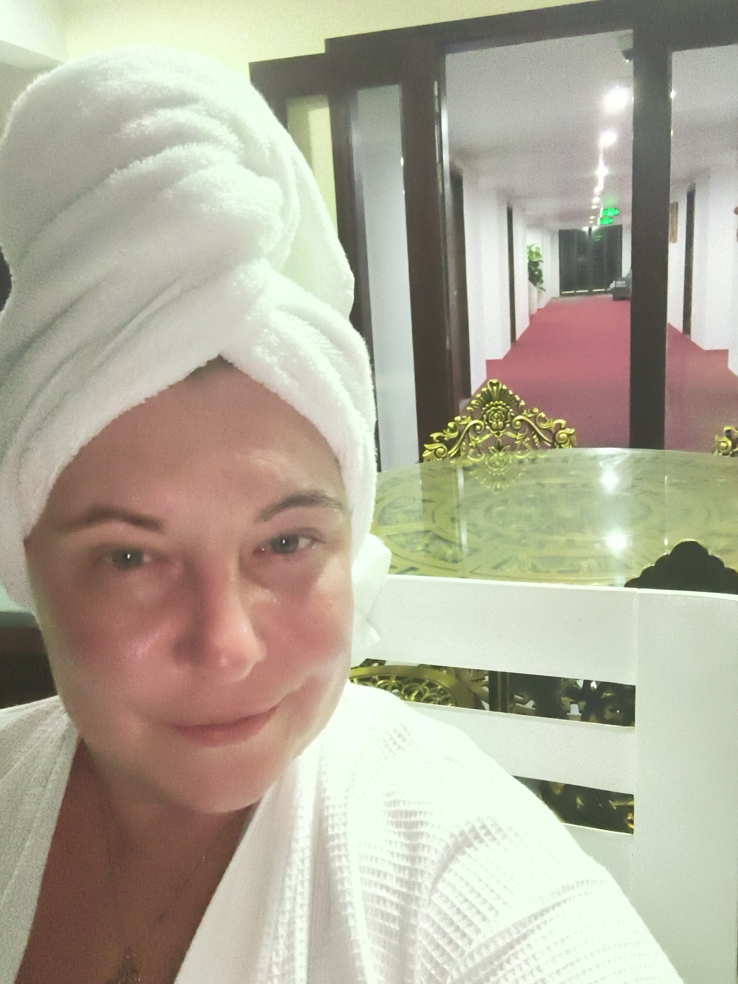A woman, with her hair wrapped in a white towel and wearing a white bathrobe, takes a selfie in what appears to be a hotel hallway or spa area. Her face is close to the camera, with her eyes open and looking towards the lens. She has light-colored skin and is smiling slightly. In the background, a long, red-carpeted hallway is visible, illuminated by overhead lights. There are doorways on either side of the hallway, and potted plants can be seen. A ornate golden table with a glass top is situated in the mid-ground. The setting suggests a resort or hotel, possibly in Phu Thuy Ward, Vietnam, given the context provided. The overall ambiance is one of relaxation and self-care.