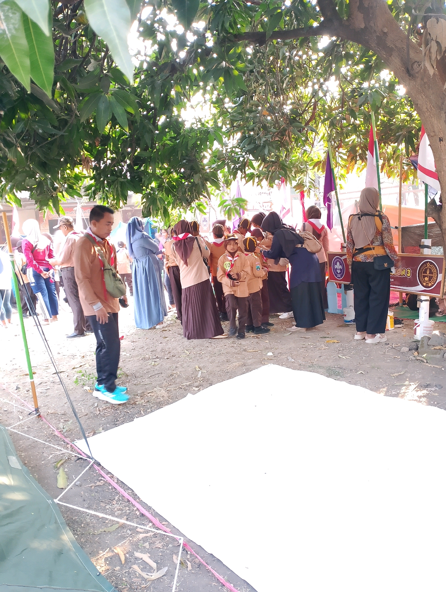 In Probolinggo, Indonesia, an outdoor event is underway beneath the dappled sunlight filtering through the leaves of a large tree. A group of people, many in scout uniforms, are gathered around a table with flags displayed prominently. Several individuals are wearing tan scout uniforms with red neckerchiefs, while others are in maroon and tan attire. A man in a tan long-sleeved shirt and black pants, wearing bright blue sneakers, stands prominently in the foreground, looking to his right. He has a light green bag slung across his chest. To his left, a white tarp or sheet is laid out on the ground, bordered by a white and pink string structure. In the background, red and white Indonesian flags are visible, along with purple flags. The ground is covered with dirt and fallen leaves. A large, dark green tent is partially visible on the left side of the frame. The overall atmosphere suggests a community gathering or activity, possibly a scouting event or educational fair.