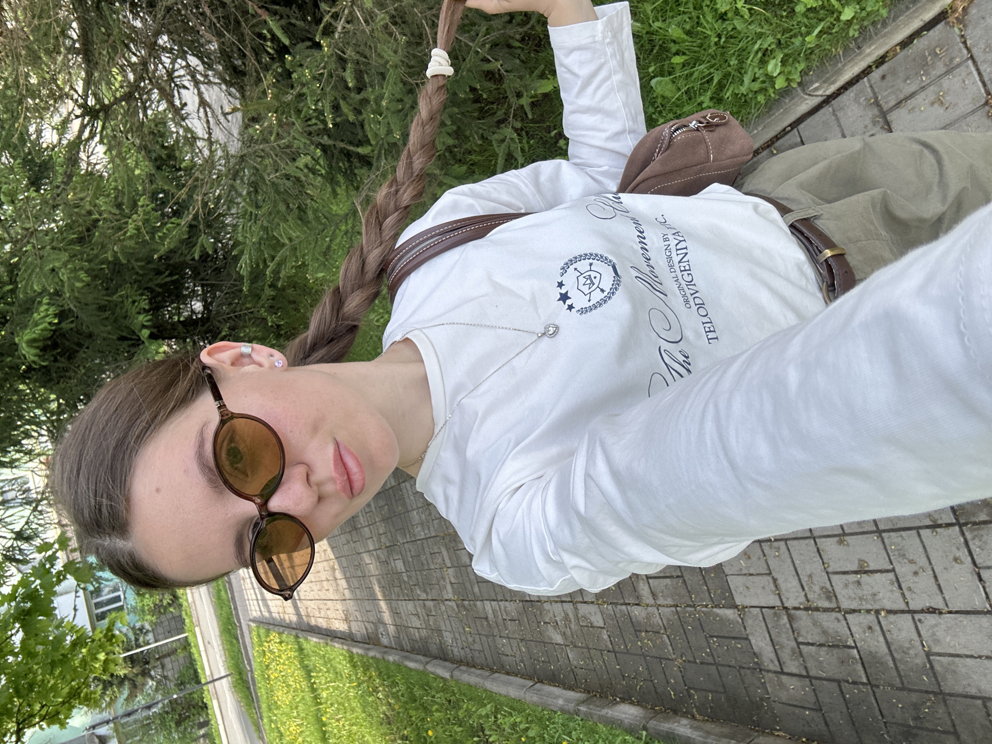 A young woman poses for a selfie outdoors. She wears round, dark-lensed sunglasses, a white long-sleeved shirt with writing and a crest, olive-green trousers, and a brown leather belt and crossbody bag. Her long brown hair is braided, held together by a white elastic. She wears earrings and a necklace with a heart pendant. The ground is paved with gray bricks and bordered by grass. In the background, there are trees and a partial view of a building. Given the context provided, the location could be Klin, Russia.