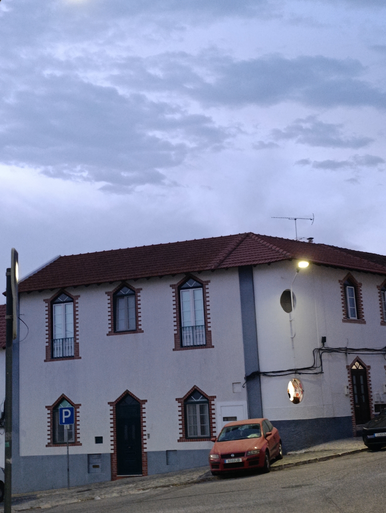 This is a low-angle, slightly blurry, exterior, full-shot photograph taken at dusk. The primary subject is a two-story building with a red tiled roof and white stucco walls accented with dark blue trim along the bottom. The building has a symmetrical facade with multiple windows. On the upper floor, from left to right, there are three arched windows with dark frames and white shutters. On the lower floor, there are two arched windows with decorative brickwork around them, one with a blue parking sign, and a dark arched doorway.
In front of the building, parked on the street, are two cars. The closest car is a red hatchback, facing the viewer, with its license plate visible. To the right, a dark-colored car is partially visible, also parked on the street. The street itself is paved and slopes upwards.
The sky above is overcast with dramatic, dark grey clouds, suggesting impending rain or twilight. A street lamp with a bright, illuminated fixture is visible on the left side of the frame, casting a warm light. A television antenna is mounted on the roof of the building on the right side. The image appears to be taken in a residential or urban setting. Based on visual cues, this location is likely in Setúbal, Portugal.