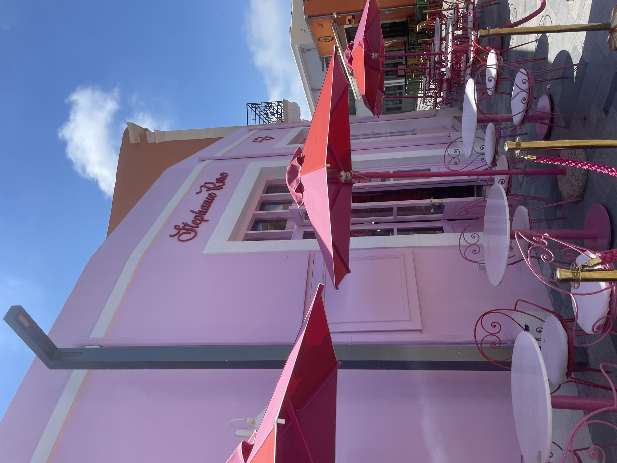 The image shows the exterior of a pink building with the name "Stephanie Rose" written in cursive script above an entrance. In front of the building, there is an outdoor seating area with several tables and chairs, some of which are pink. Red and pink umbrellas provide shade over some of the tables. The building is located in Paphos Municipality, Cyprus. The area appears to be a commercial street or a tourist area, with other buildings visible in the background, one of which is orange. The sky is blue with some white clouds, indicating it is daytime. There are no people visible in the immediate seating area.