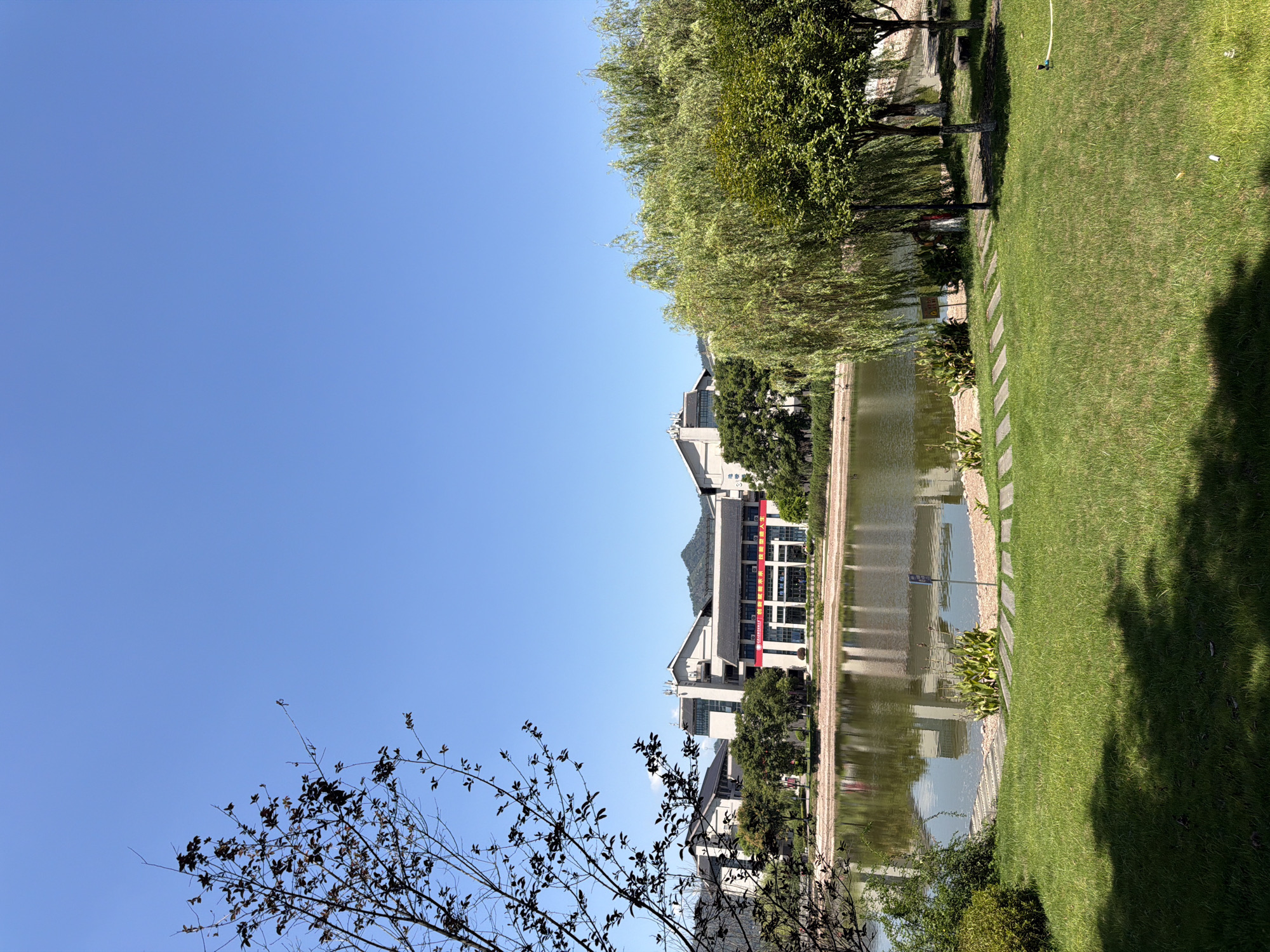 The image depicts a tranquil scene at the campus of Chongqing University of Science and Technology in Jiangjin District, Chongqing, China. In the foreground, a lush green lawn with dappled sunlight creates a serene atmosphere. A pathway of stepping stones leads towards a tranquil lake. The lake's surface reflects the clear blue sky and the surrounding buildings. Across the lake, modern buildings with traditional Chinese architectural elements stand as a testament to the institution's blend of heritage and modernity. Some buildings feature red banners with Chinese characters, indicating an event or announcement. In the background, a range of mountains adds a natural backdrop to the cityscape. Lush green trees, including weeping willows, frame the scene, contributing to the peaceful ambiance. The image is captured on a bright, sunny day, highlighting the vibrant colors of the environment. There are no visible people in the image.