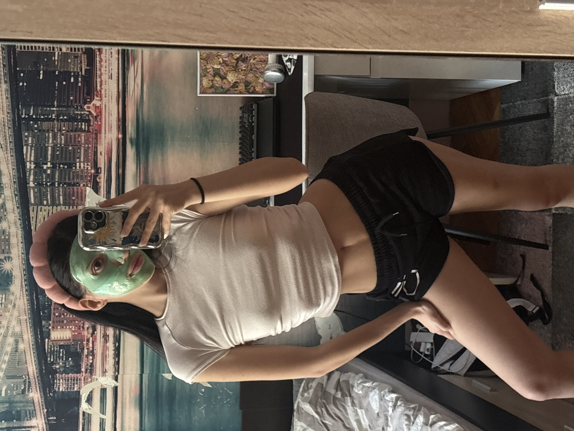 A person with a green face mask is taking a selfie in a mirror. The person is wearing a pink headband, a white short-sleeved shirt, and black shorts. The background includes a city skyline mural, a picture frame, a desk, a chair, and a bed. The location appears to be indoors, possibly a bedroom. The city is unknown.