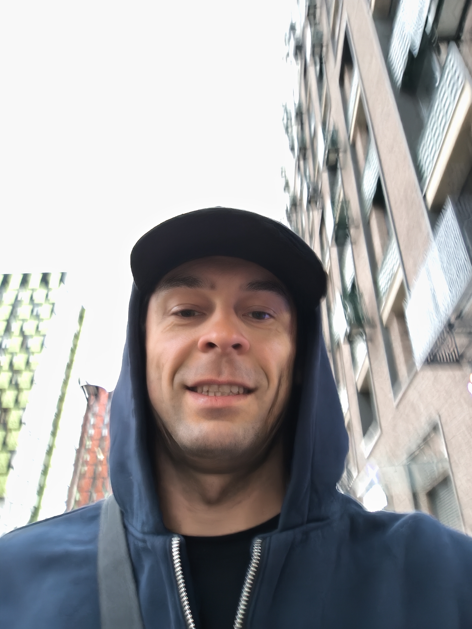 A selfie captured outdoors depicts a man wearing a dark-colored baseball cap and a blue hooded jacket with a silver zipper. The background shows urban buildings, one with green and white structures and another taller building with windows. The man is looking directly at the camera, smiling slightly. The location appears to be an urban area; however, definitive proof of Moscow, Russia, cannot be confirmed solely from this image.